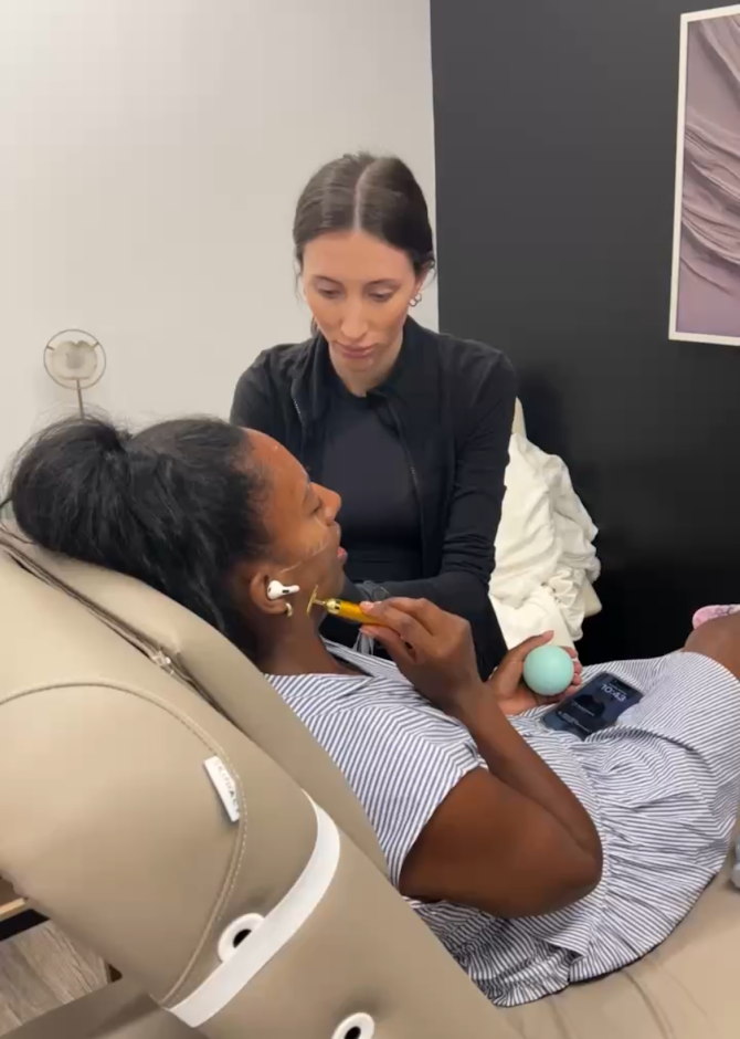 A woman lying in a medical chair receiving a procedure from a healthcare worker standing beside her, in a clinical setting.