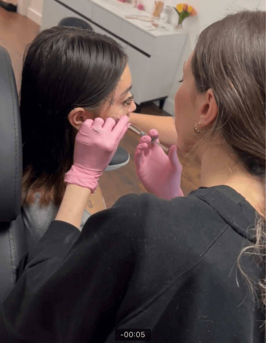 Two women wearing pink gloves are in a close-up, one applying a cosmetic or medical treatment to the other's face.