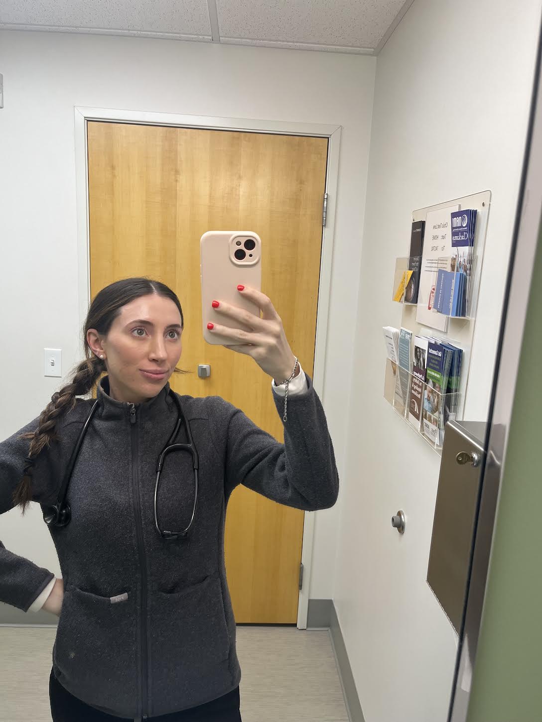 Woman taking a selfie in a mirror, holding phone with right hand, standing in a room with a wooden door and wall-mounted items.