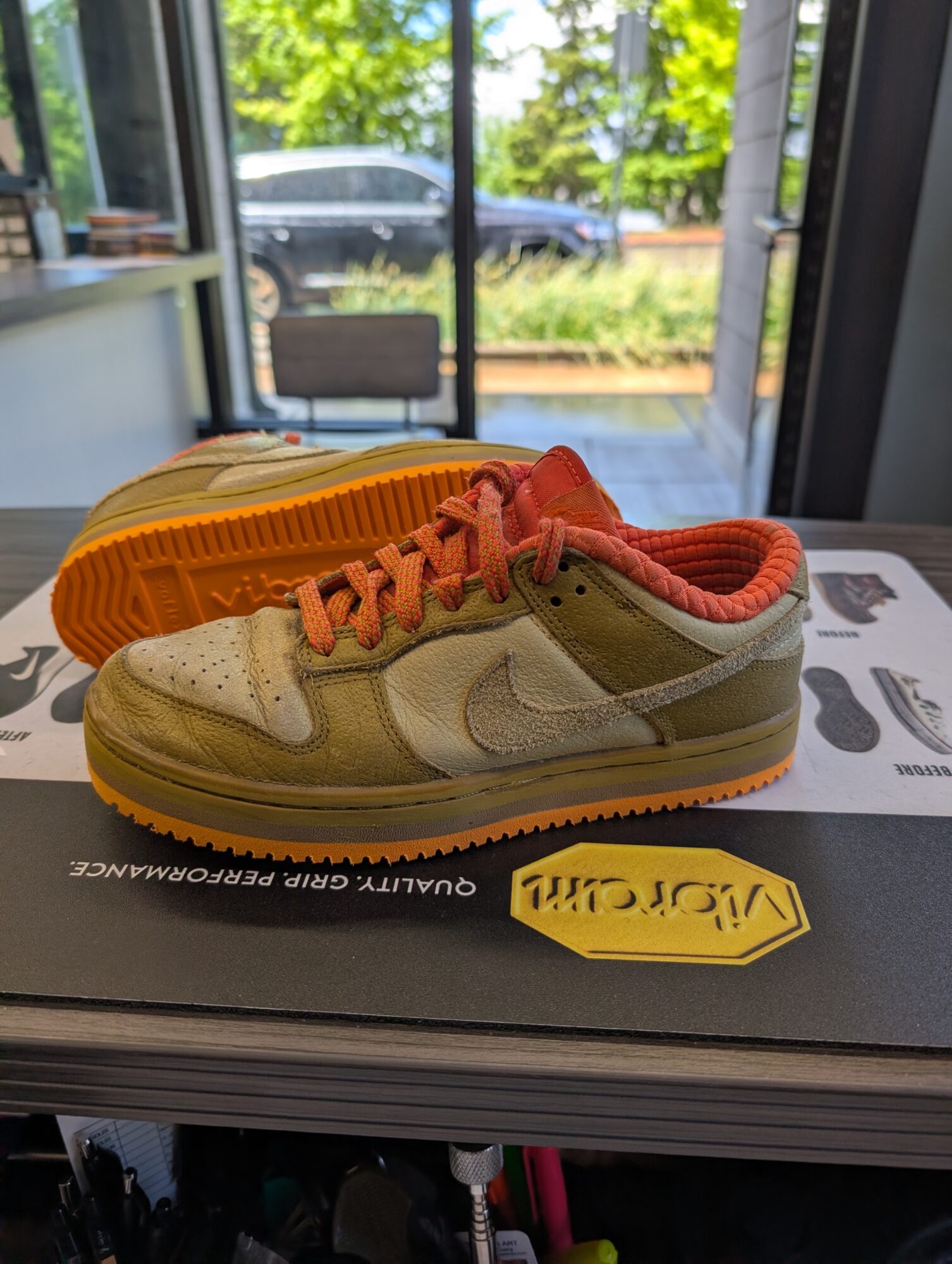 A sneaker with beige, orange, and yellow colors on a black box, with a glass door and outdoor scene in background.