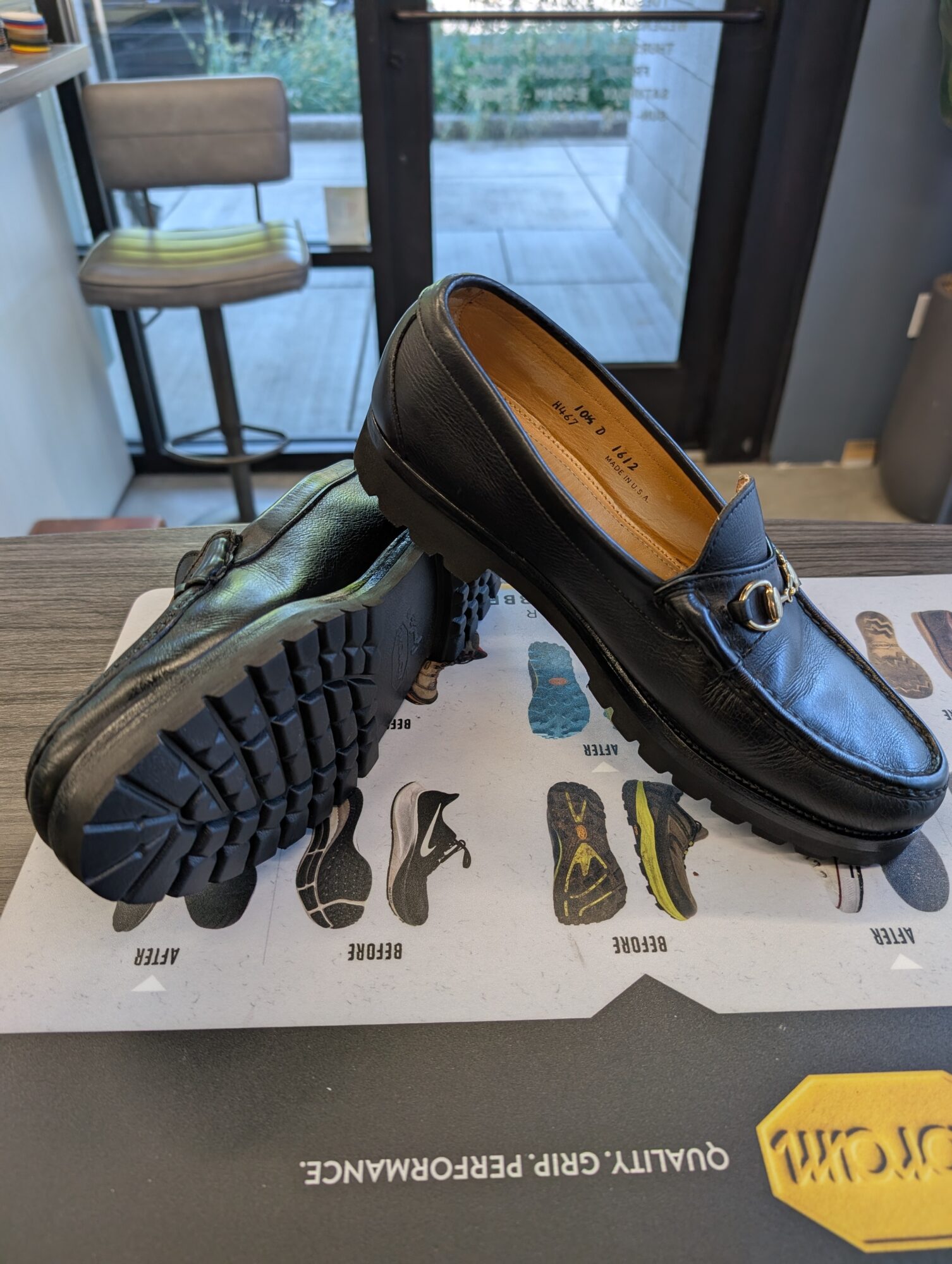 Pair of black loafers on a table, one shoe upright and the other tilted, with a window and outdoor view in background.