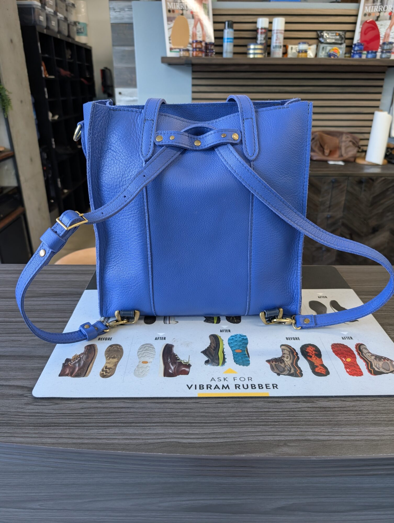 Blue handbag with a strap, placed on a counter with shoe samples and a sign reading 'VIBRAM RUBBER'.