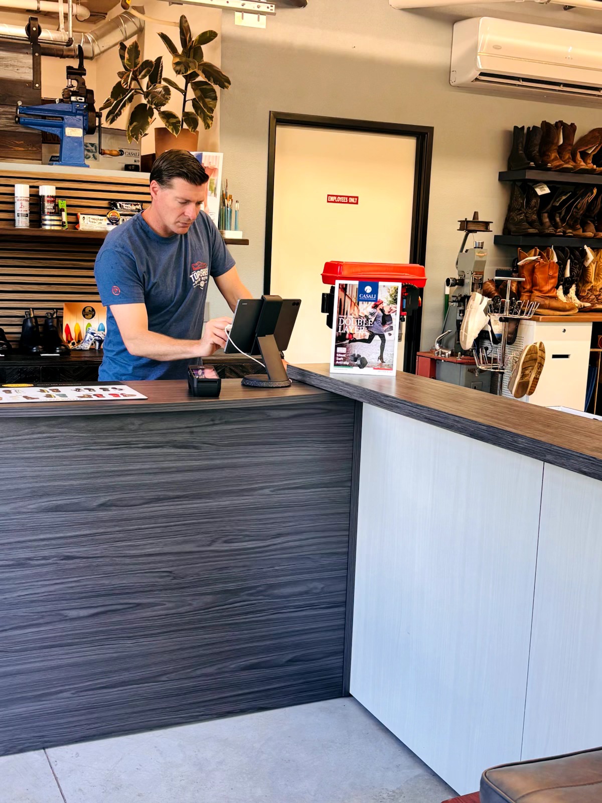 A man stands behind a counter in a store, with shelves of shoes and plants in the background.