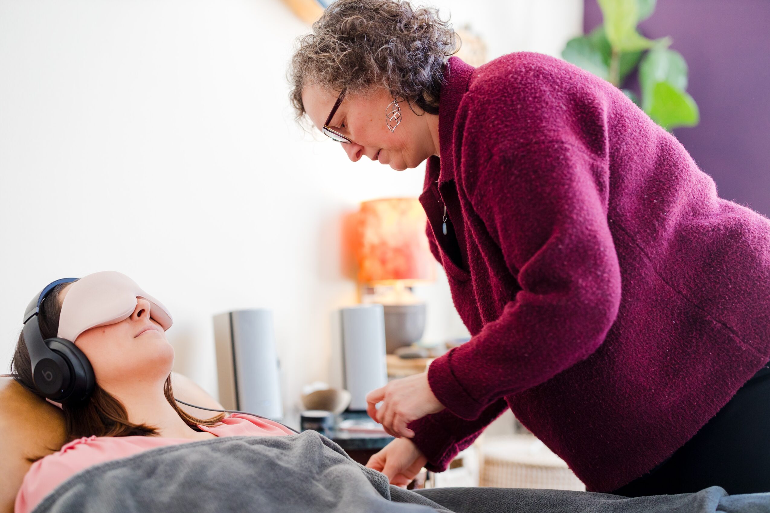 Older woman leaning over a young girl lying on a bed, listening to music with headphones, in a bright room.