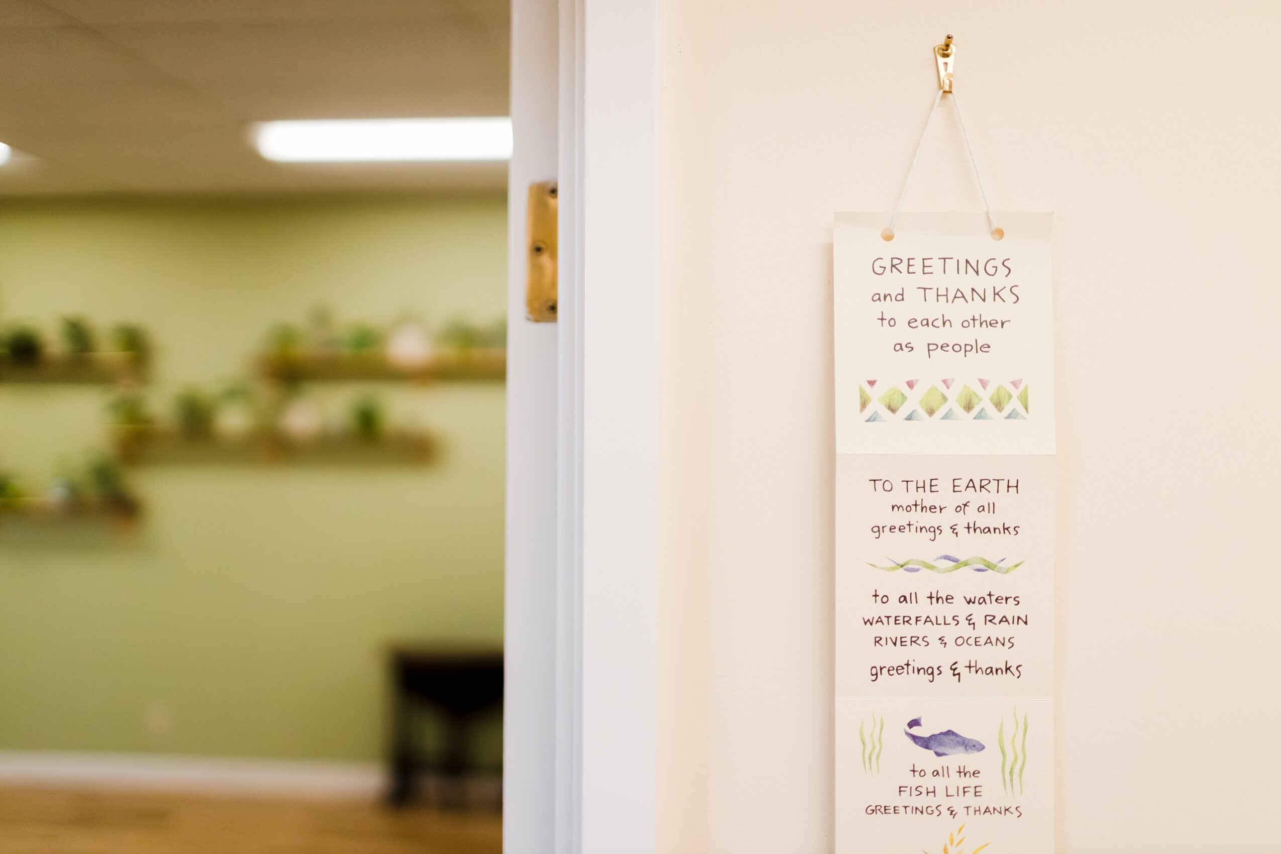 Vertical wall hanging with handwritten messages and small illustrations, hanging on a white wall, with a blurred room in the background.