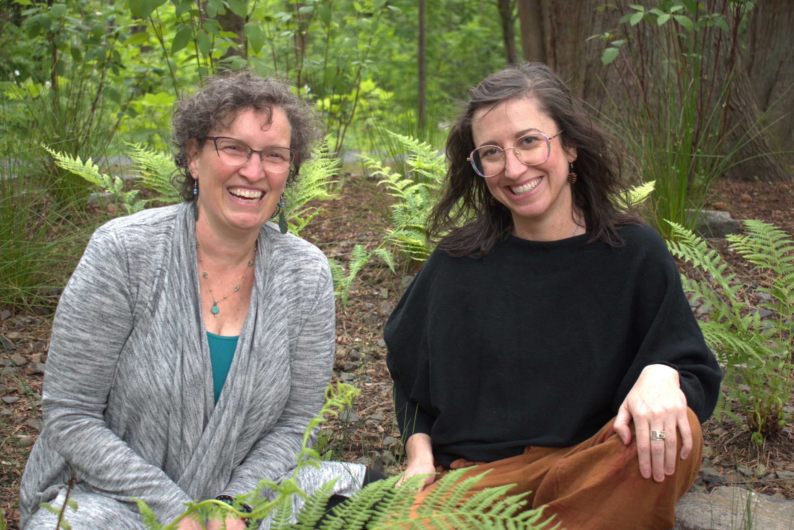 Two women smiling outdoors in a lush green setting, one wearing a gray jacket and the other a black top.