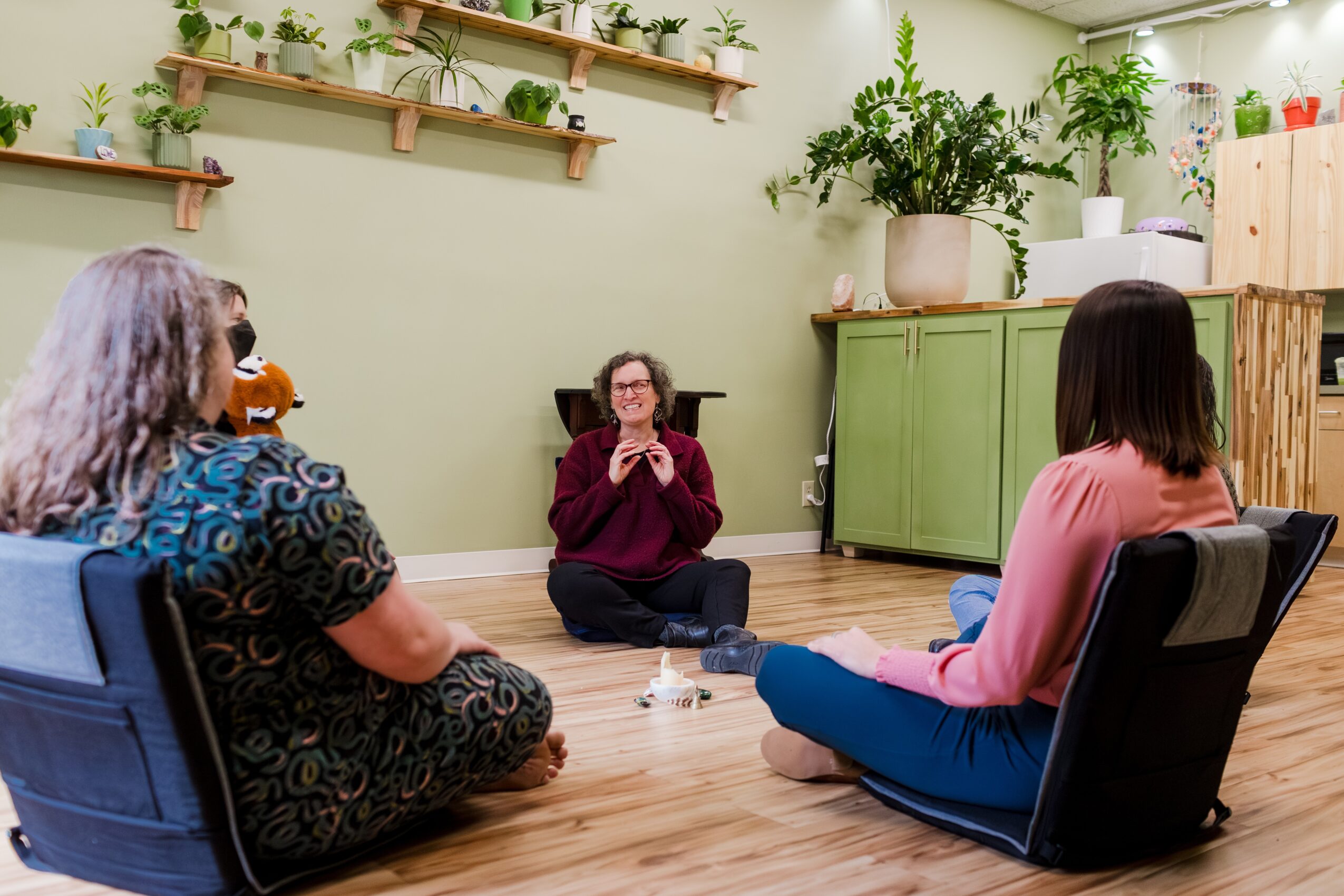 Three women sitting on the floor and in chairs in a room with green cabinets and shelves with plants.