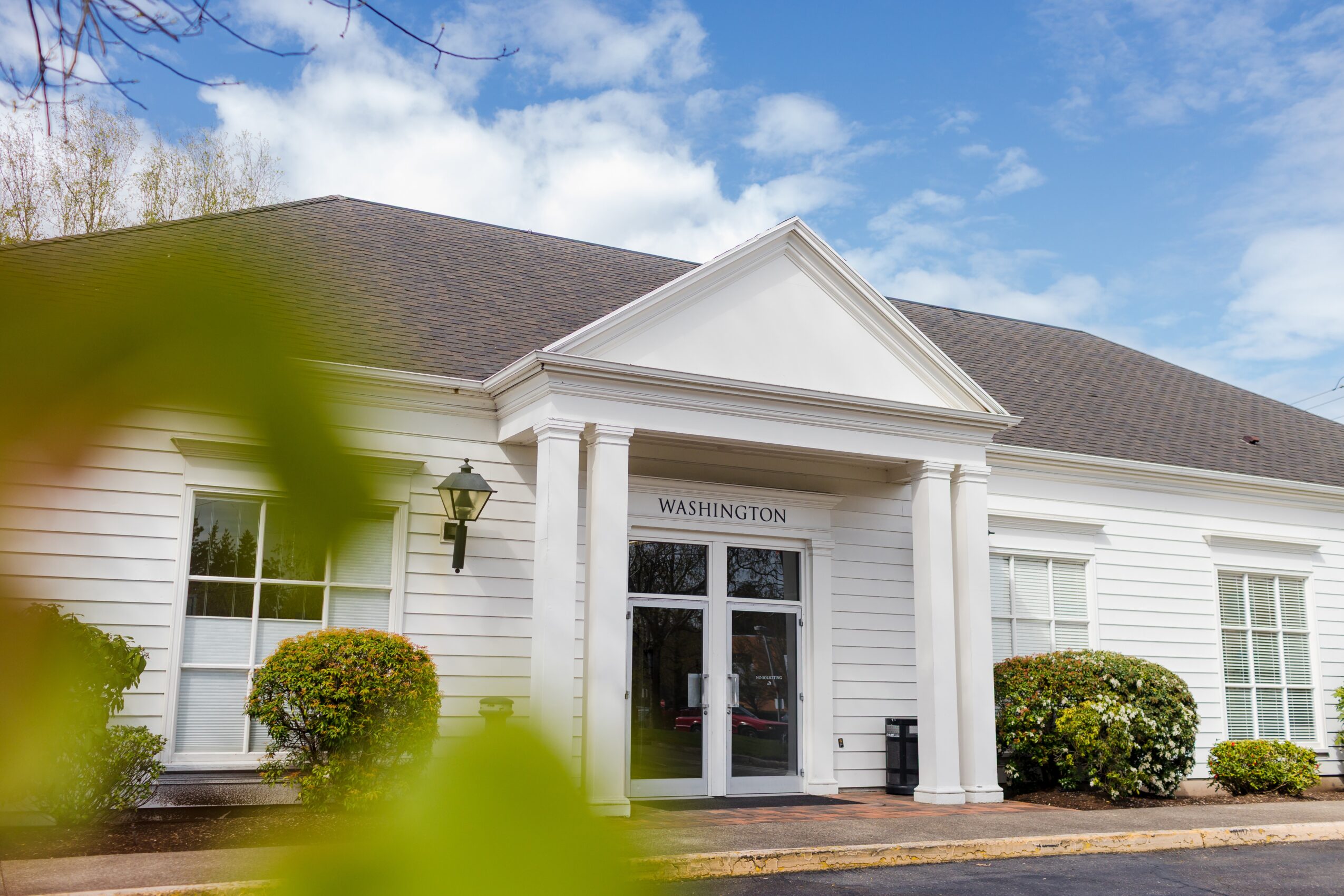 White building with a gabled roof, entrance with columns, and bushes in front, under a partly cloudy sky.