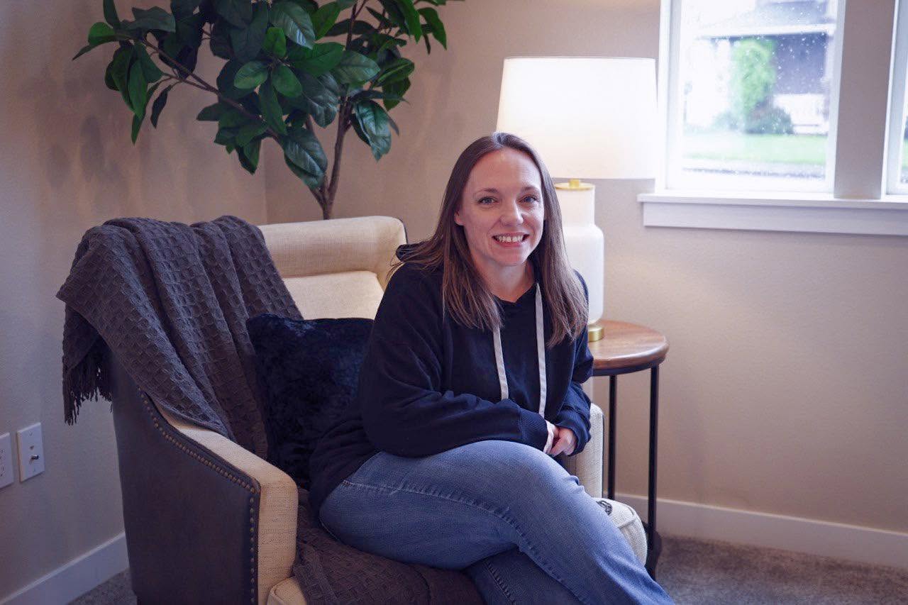 Woman sitting on a beige armchair near a window, smiling, with a blanket draped over the chair's back, in a cozy room.