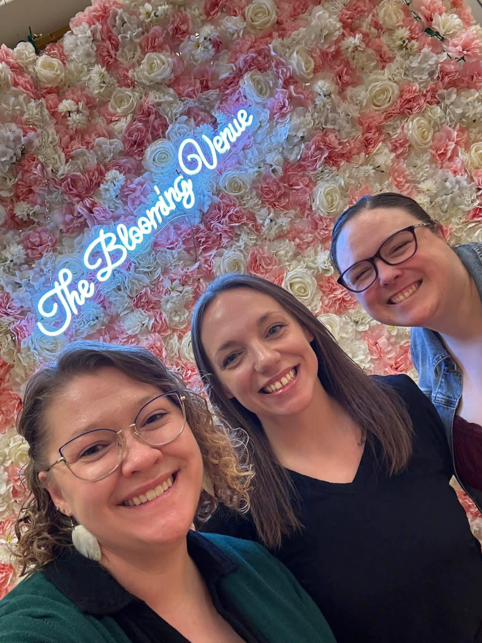 Three women smiling in front of a floral wall with a neon sign that reads 'The Blooming Venue'.