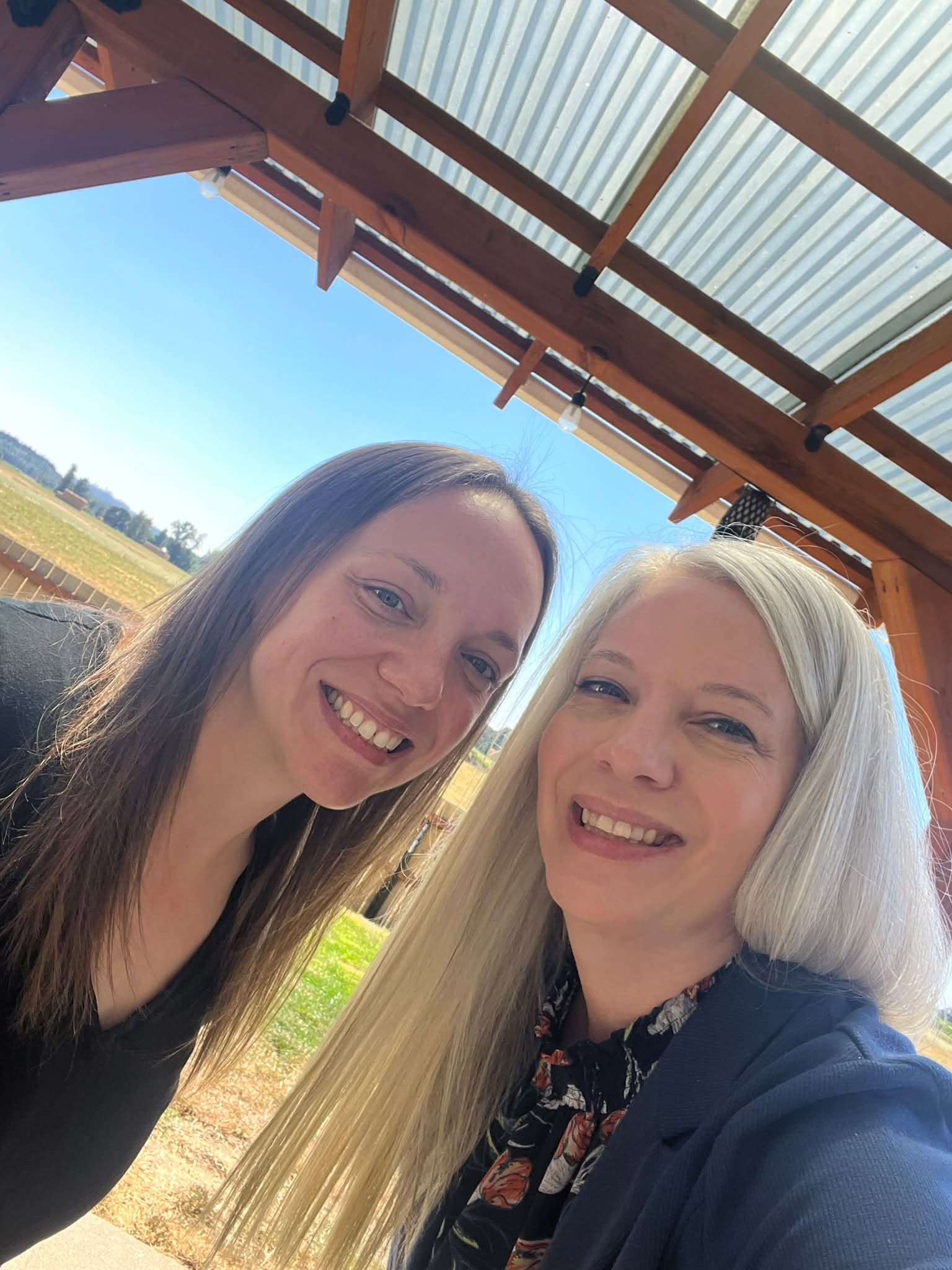 Two women smiling outdoors under a wooden and metal roof structure, with a field and blue sky in the background.