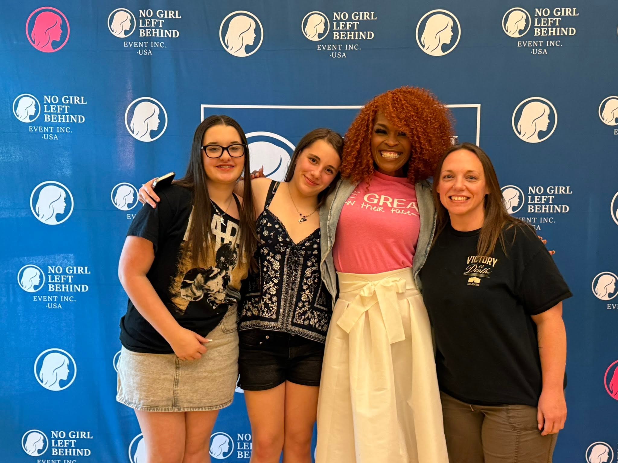 Four women standing together in front of a blue backdrop with logos, smiling for the photo.