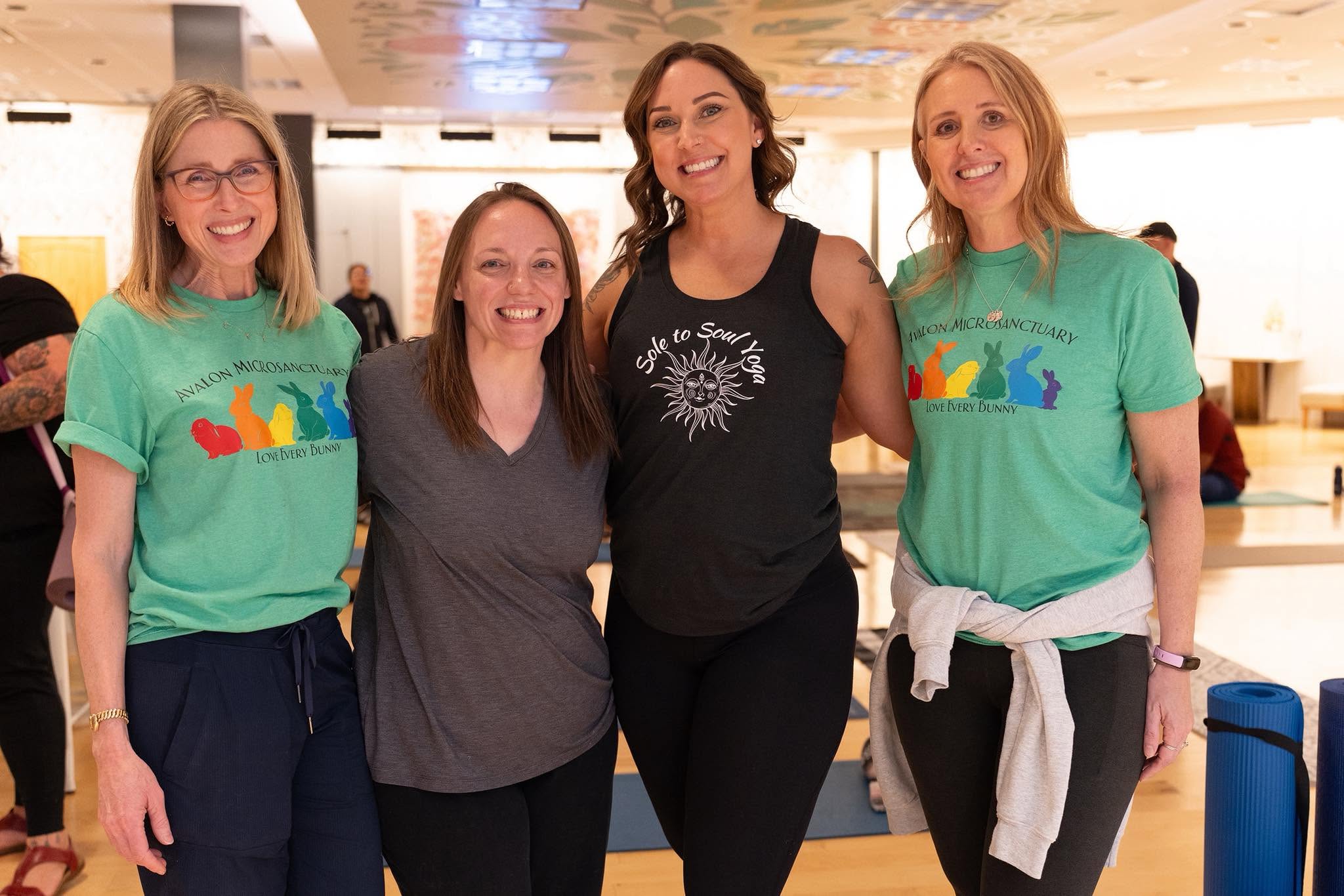 Four women standing together indoors, smiling, with a bright background and casual clothing.