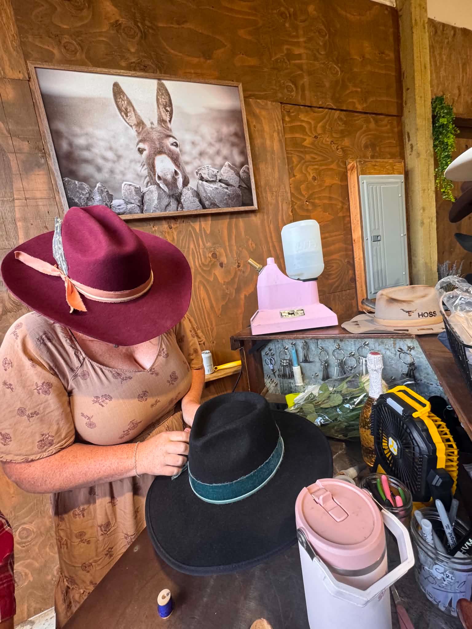 Woman wearing a pink hat and floral shirt looking at a black hat on a table, with a framed animal photo on the wall behind.