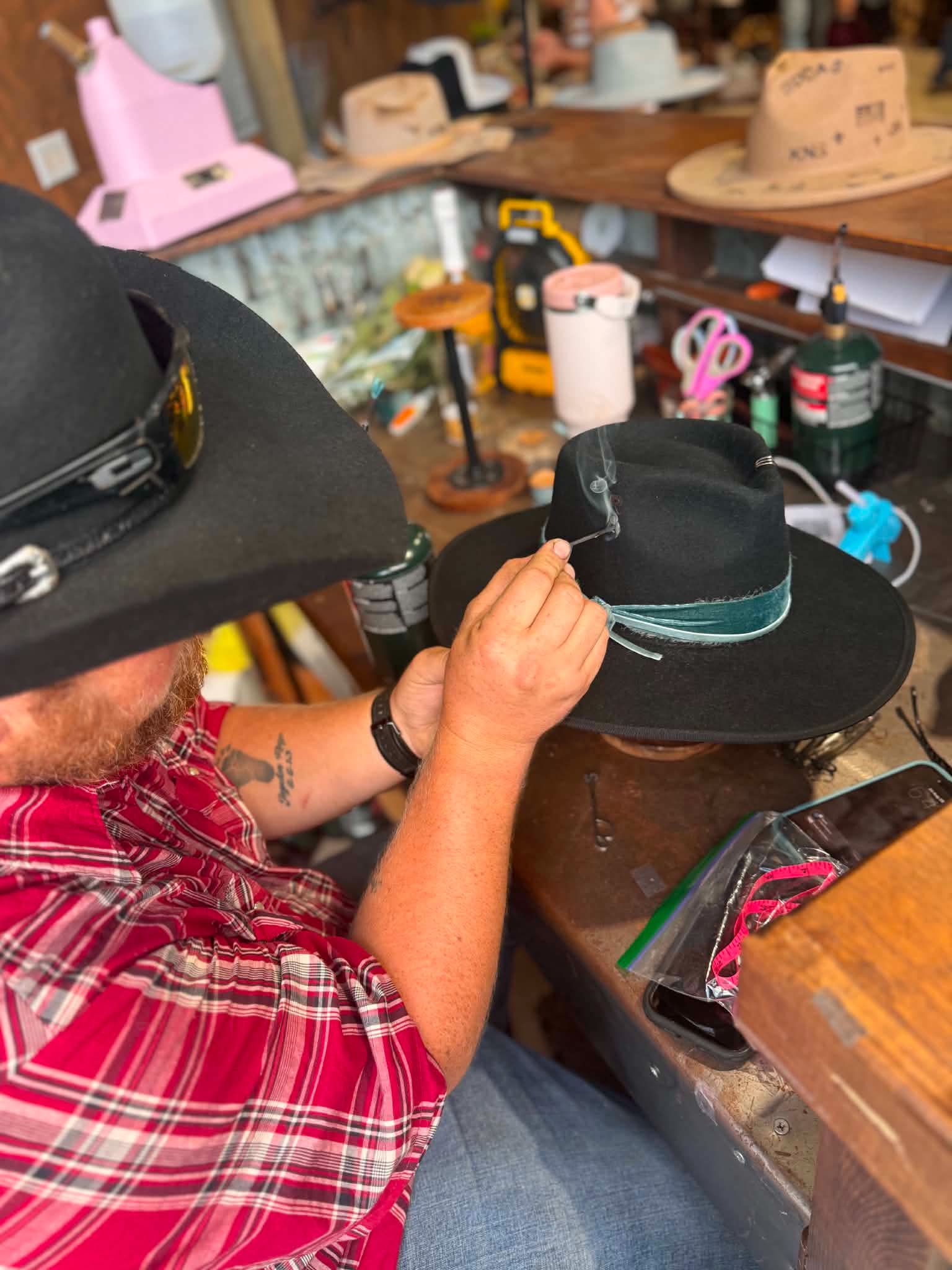 Person in a red plaid shirt and black hat working on a black hat at a cluttered workbench.