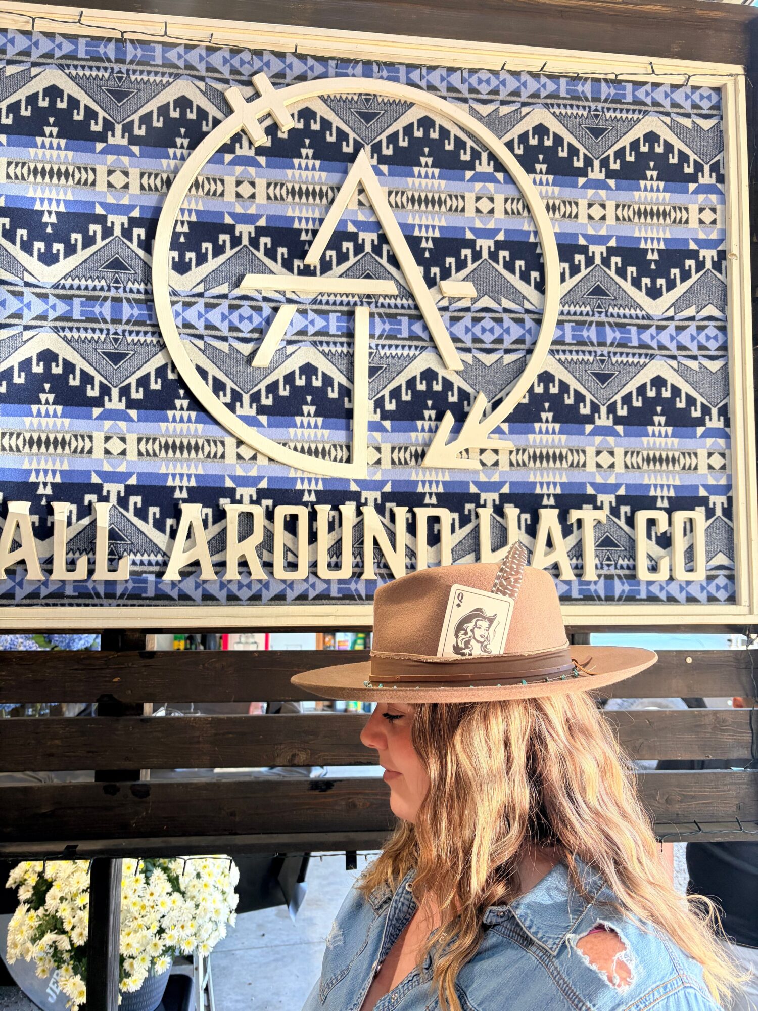 Person with long hair wearing a hat and denim jacket, facing a decorative wall with geometric patterns and text.