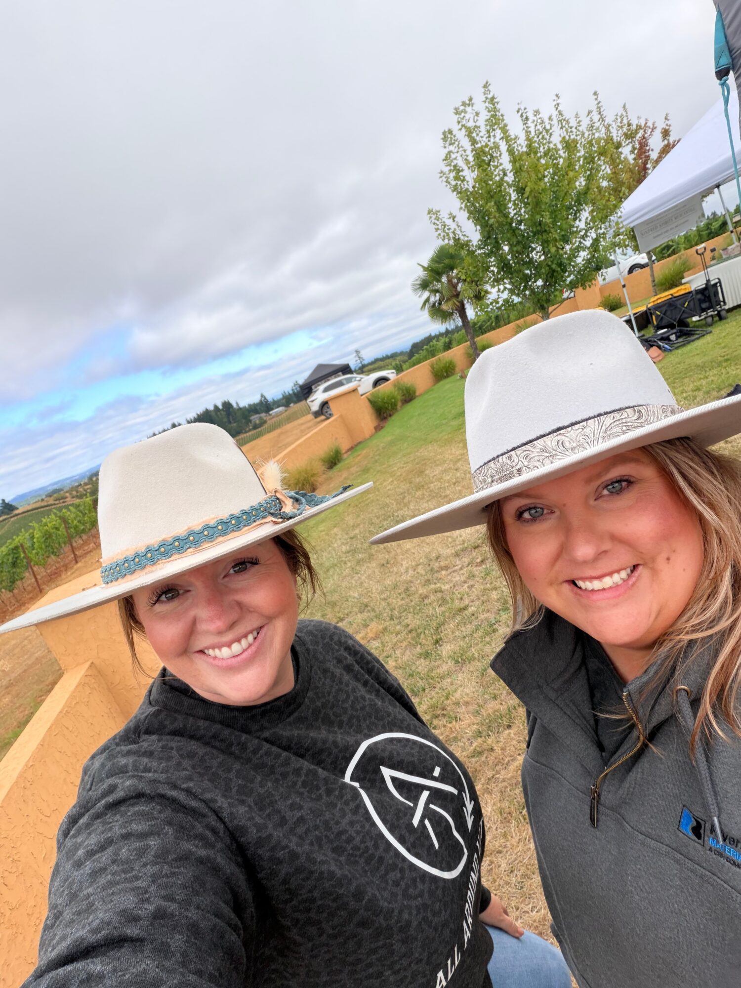Two women wearing white hats smiling outdoors with a fence, trees, and cloudy sky in the background.
