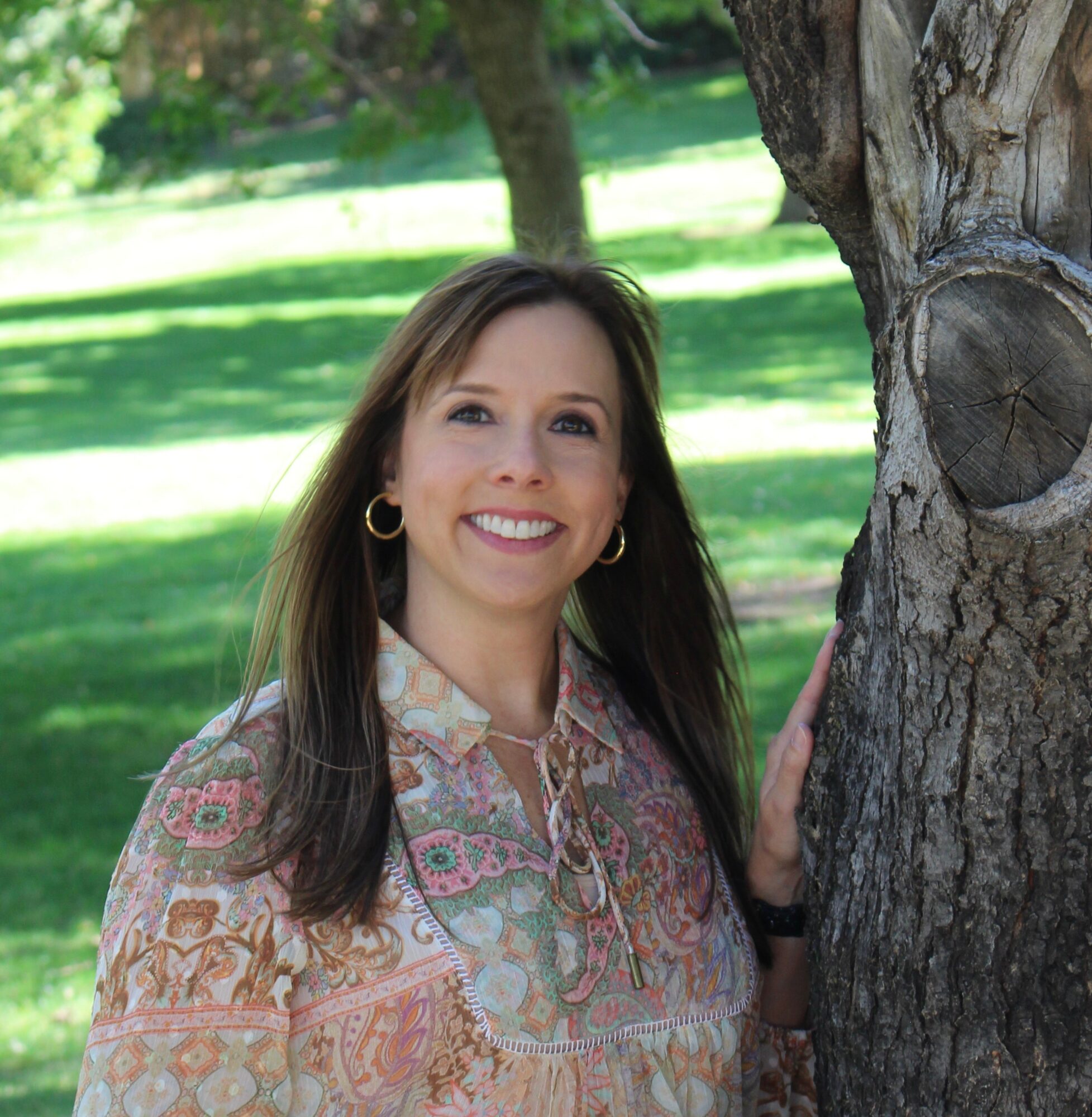 Smiling woman with long brown hair, wearing a patterned blouse, standing outdoors near a tree in a park.