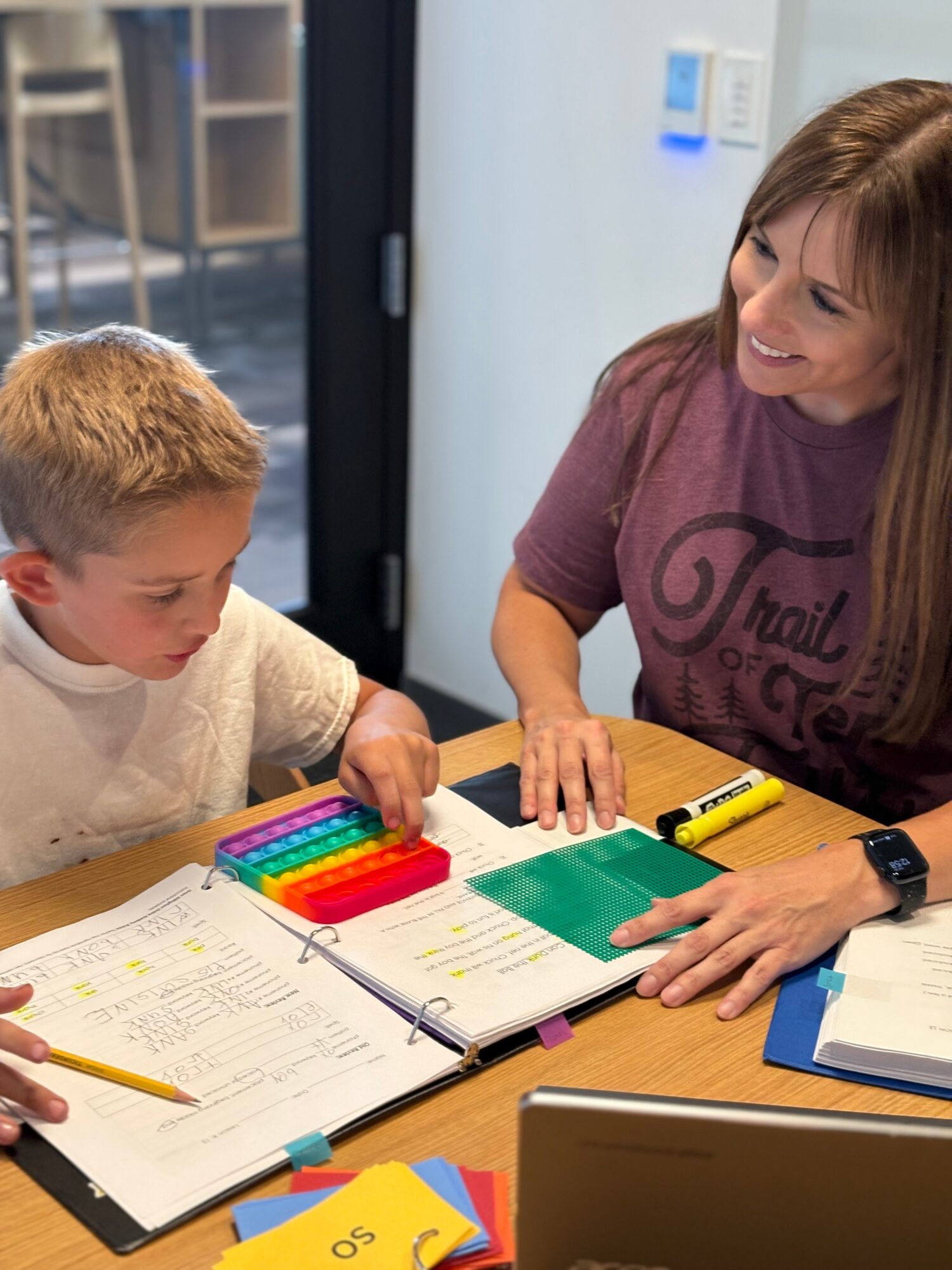 A woman and a young boy sit at a table with books, papers, and colorful markers, engaging in a learning activity.