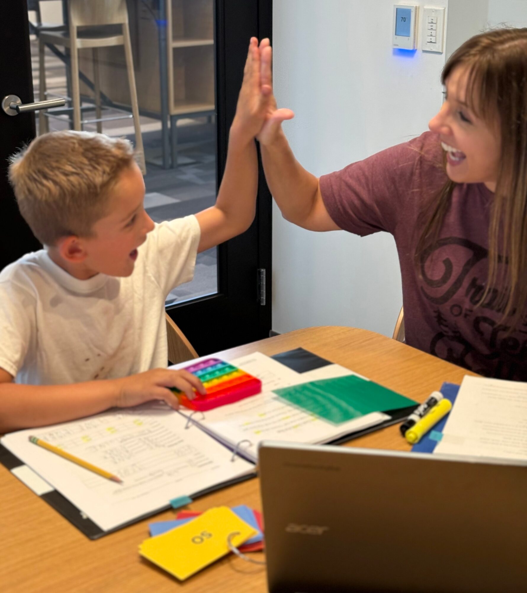 Child and adult high-fiving at a table with books, papers, and a laptop, smiling and engaging in activity.