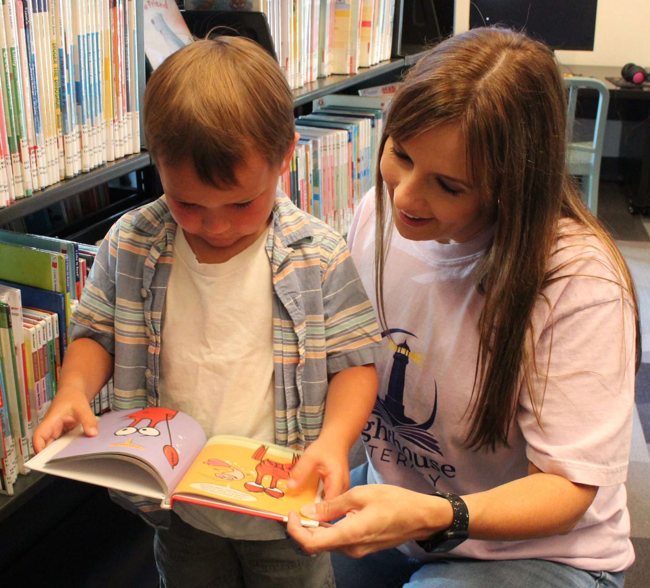 A young boy and a woman look at a book together in a library, with bookshelves in the background.