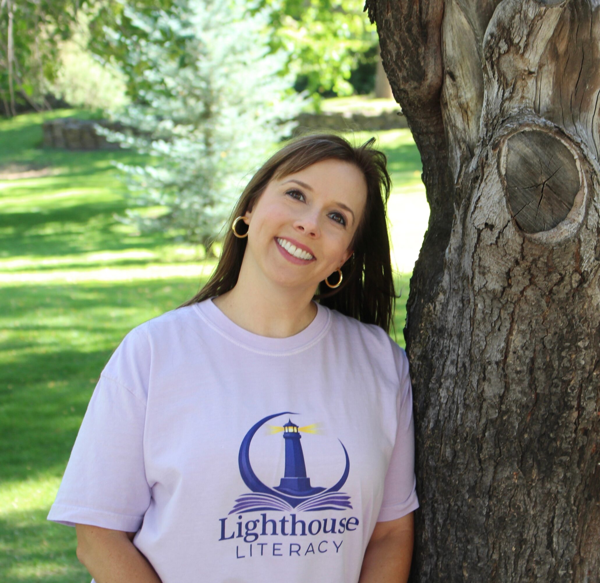 Smiling woman with dark hair and hoop earrings stands outdoors next to a tree, wearing a Lighthouse Literacy T-shirt.