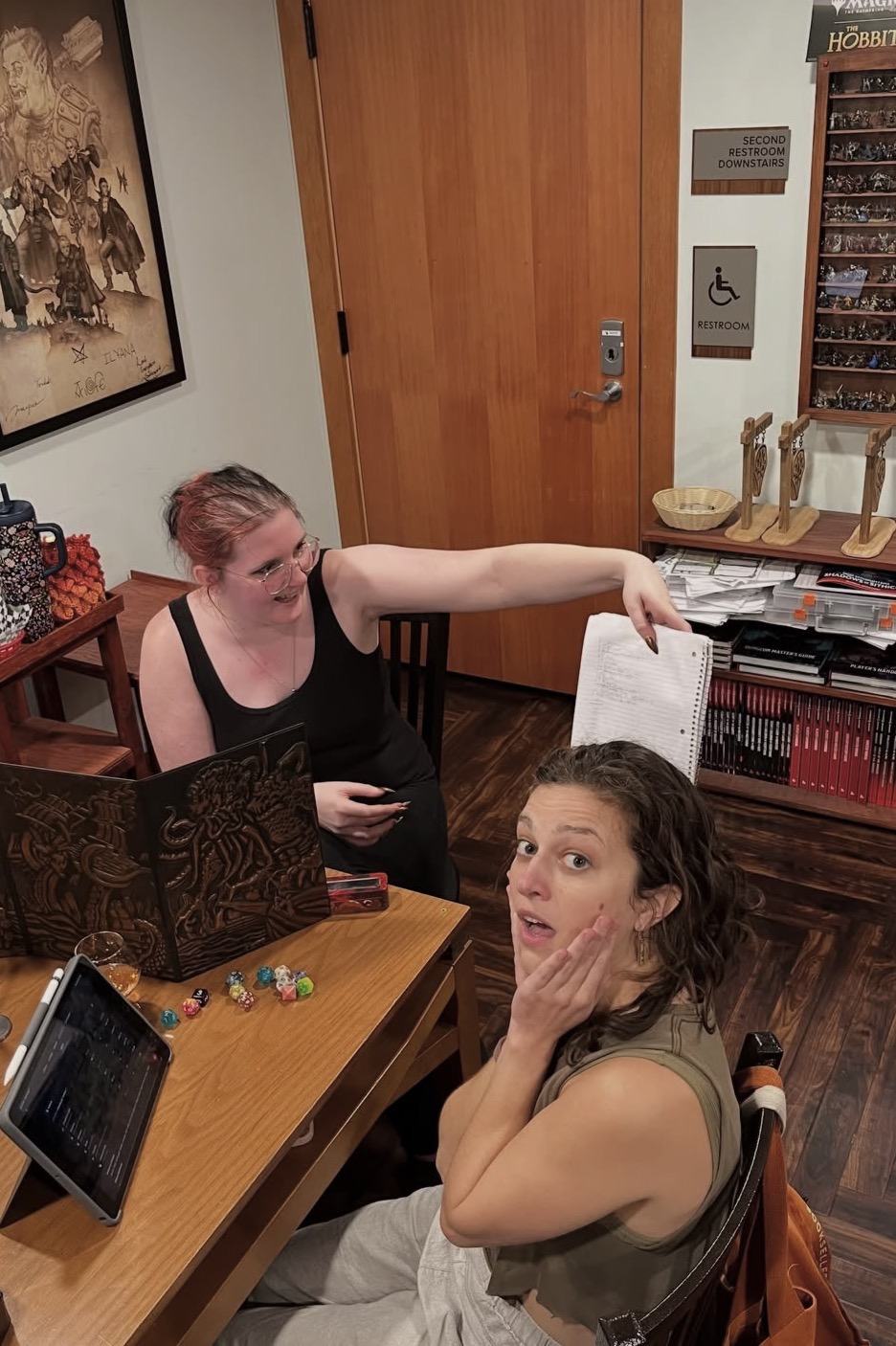 Two women at a desk, one pointing at a computer screen, the other with hand on face, in a room with wooden door and shelves.
