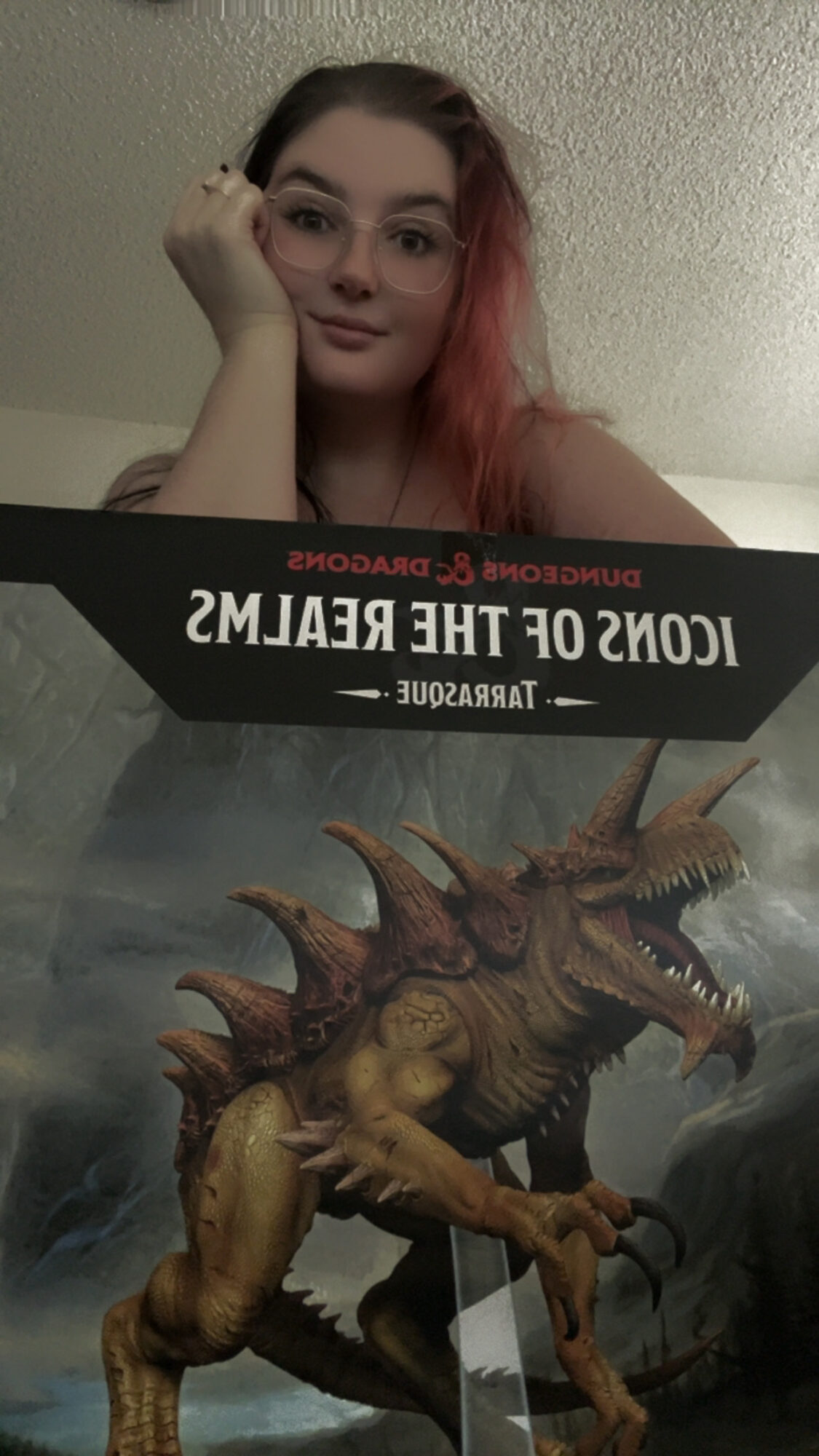 Young woman with pink hair resting her head on her hand, smiling, with a dinosaur poster in front of her.