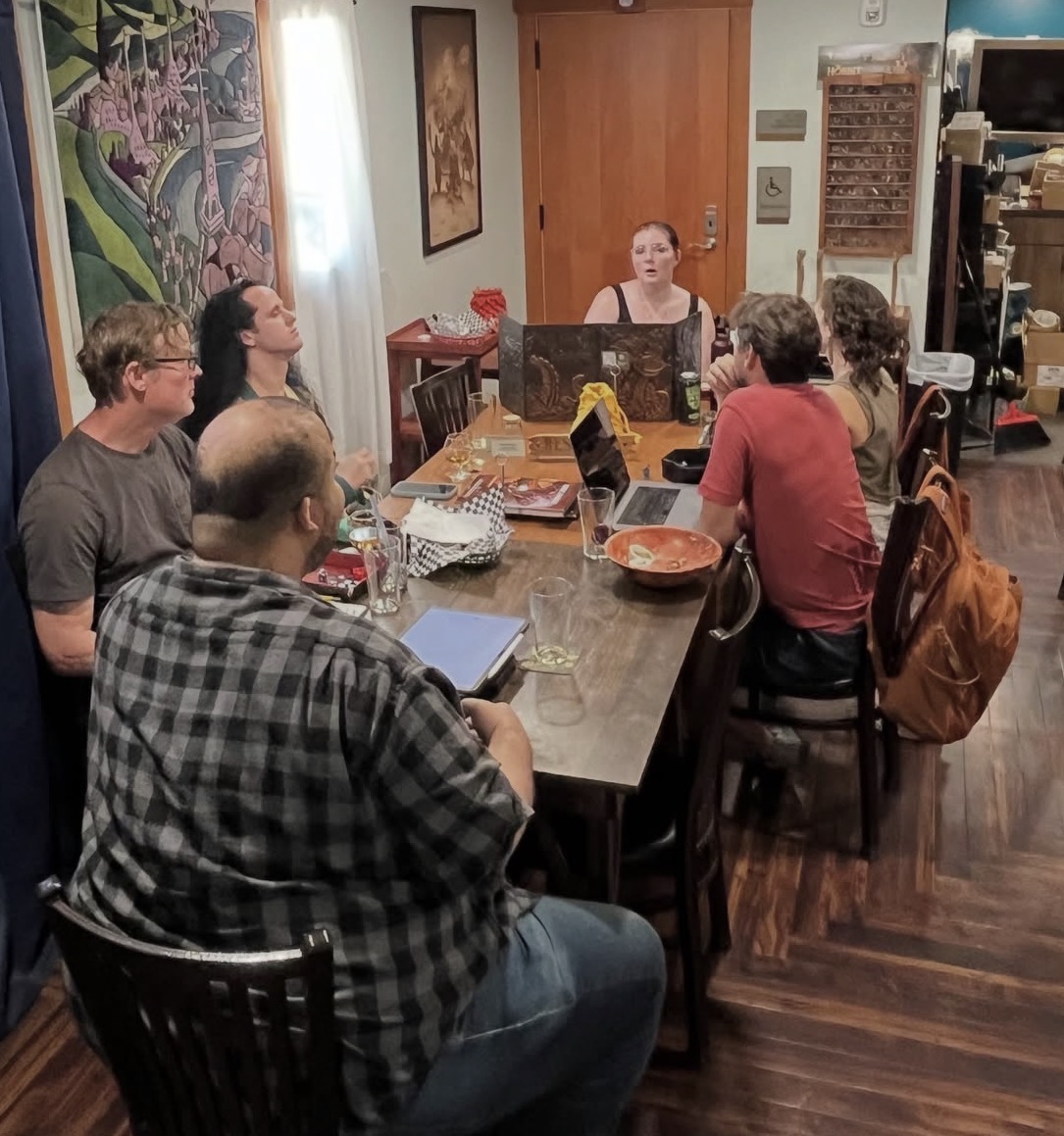 Six people sit around a dining table in a room with artwork and a door, engaged in conversation. There are laptops and bowls on the table.