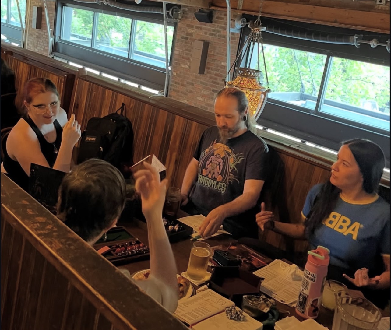 Group of four people sitting at a table in a restaurant, engaging in conversation, with windows and wooden decor in background.