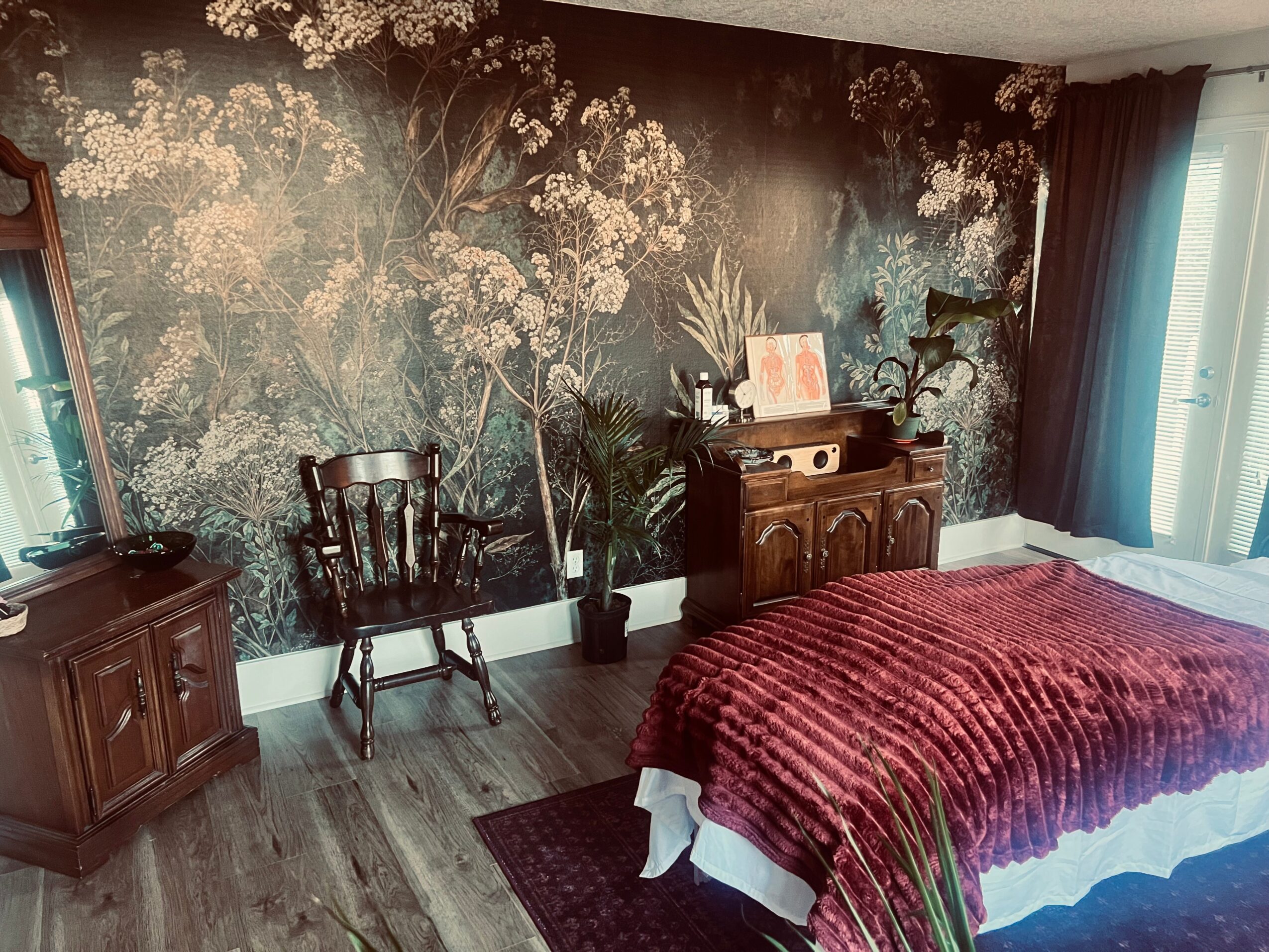Bedroom with dark floral wallpaper, wooden furniture, a bed with a red blanket, and a window with curtains.