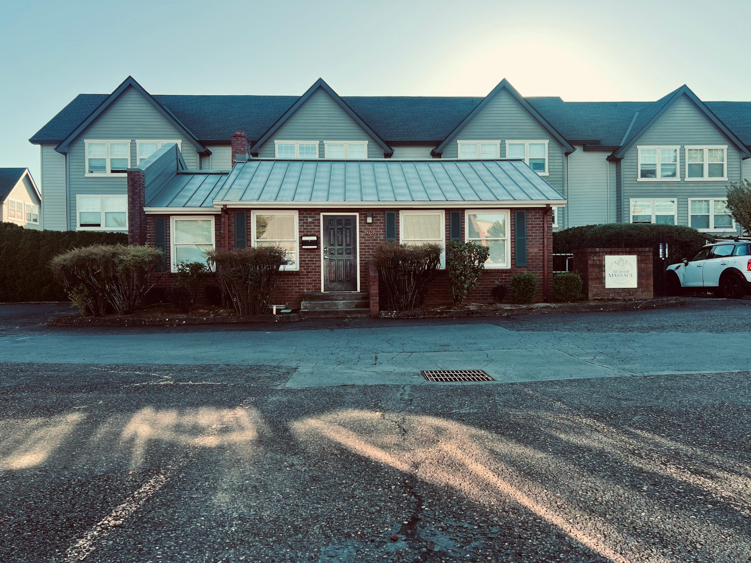 Small brick building with a metal roof, bushes in front, and a parking lot with cars, in front of larger residential buildings.