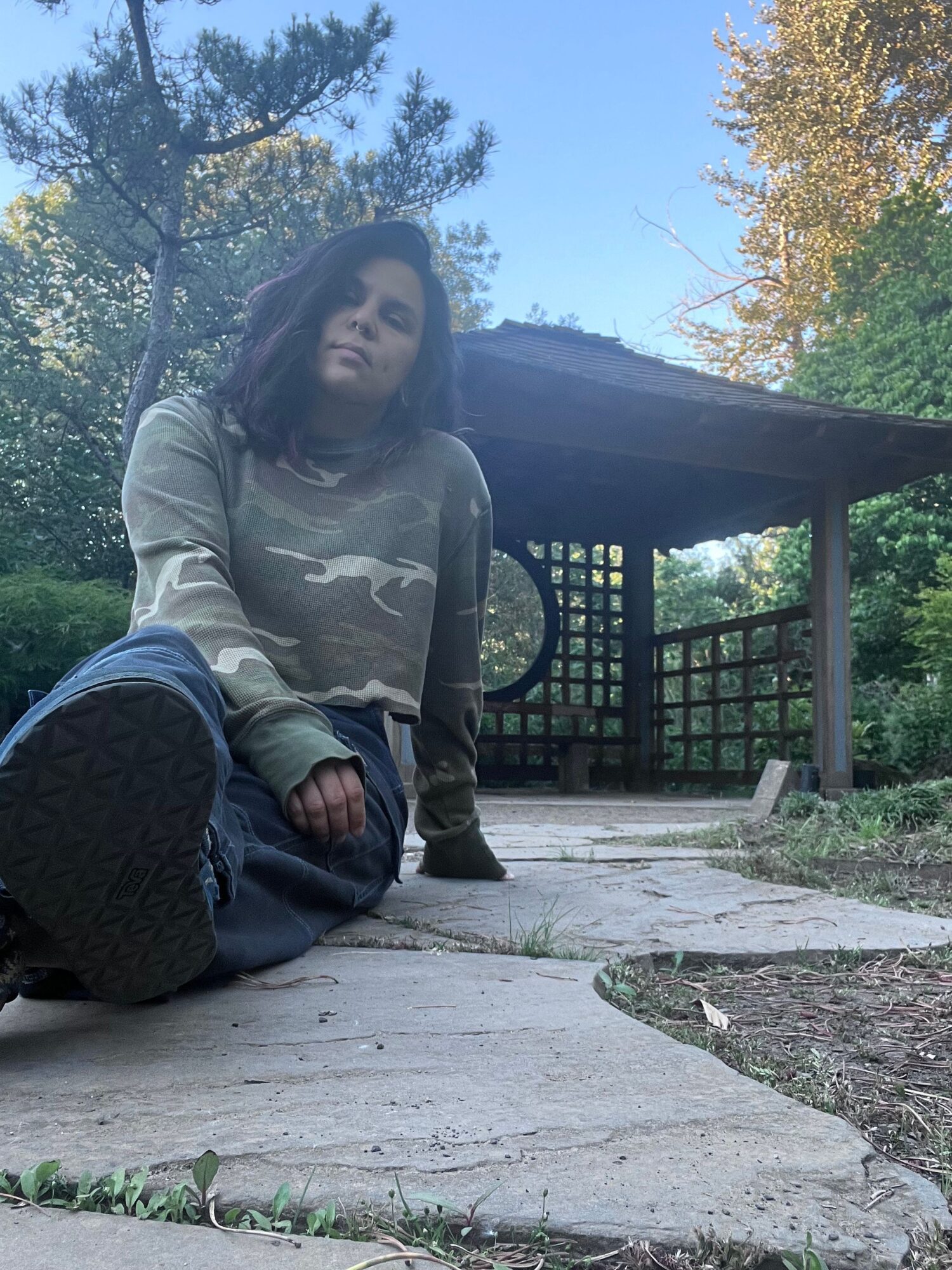 Person sitting on a stone step outdoors, wearing a light-colored hoodie and dark pants, with trees and a wooden structure in the background.