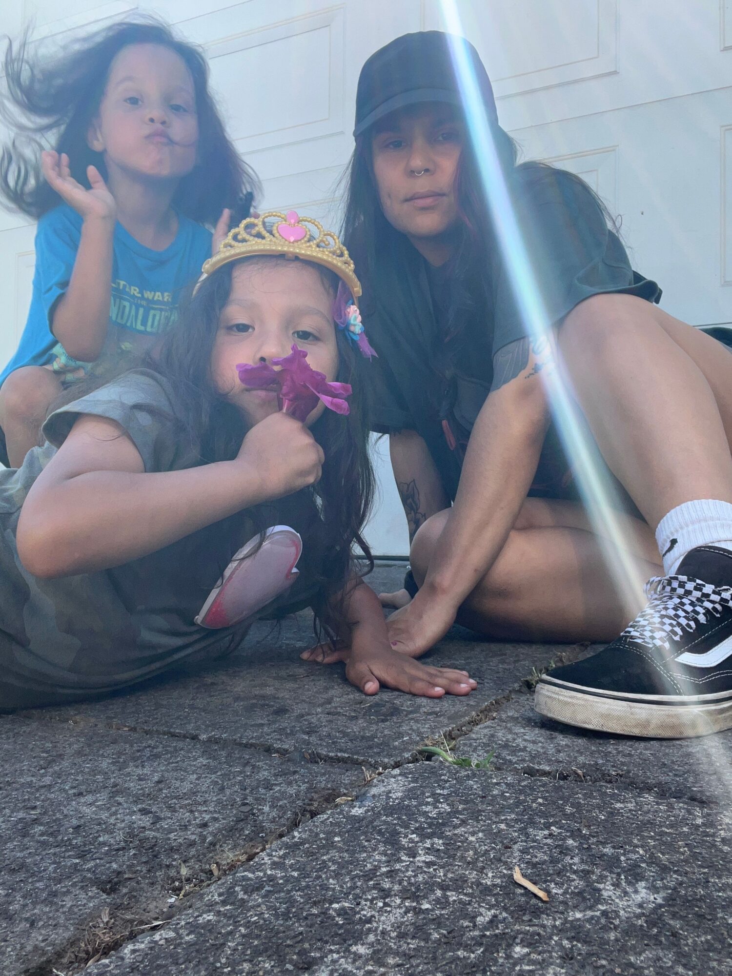 Two children sitting on a sidewalk, one wearing a crown and holding a purple object, the other making a peace sign, with an adult nearby.