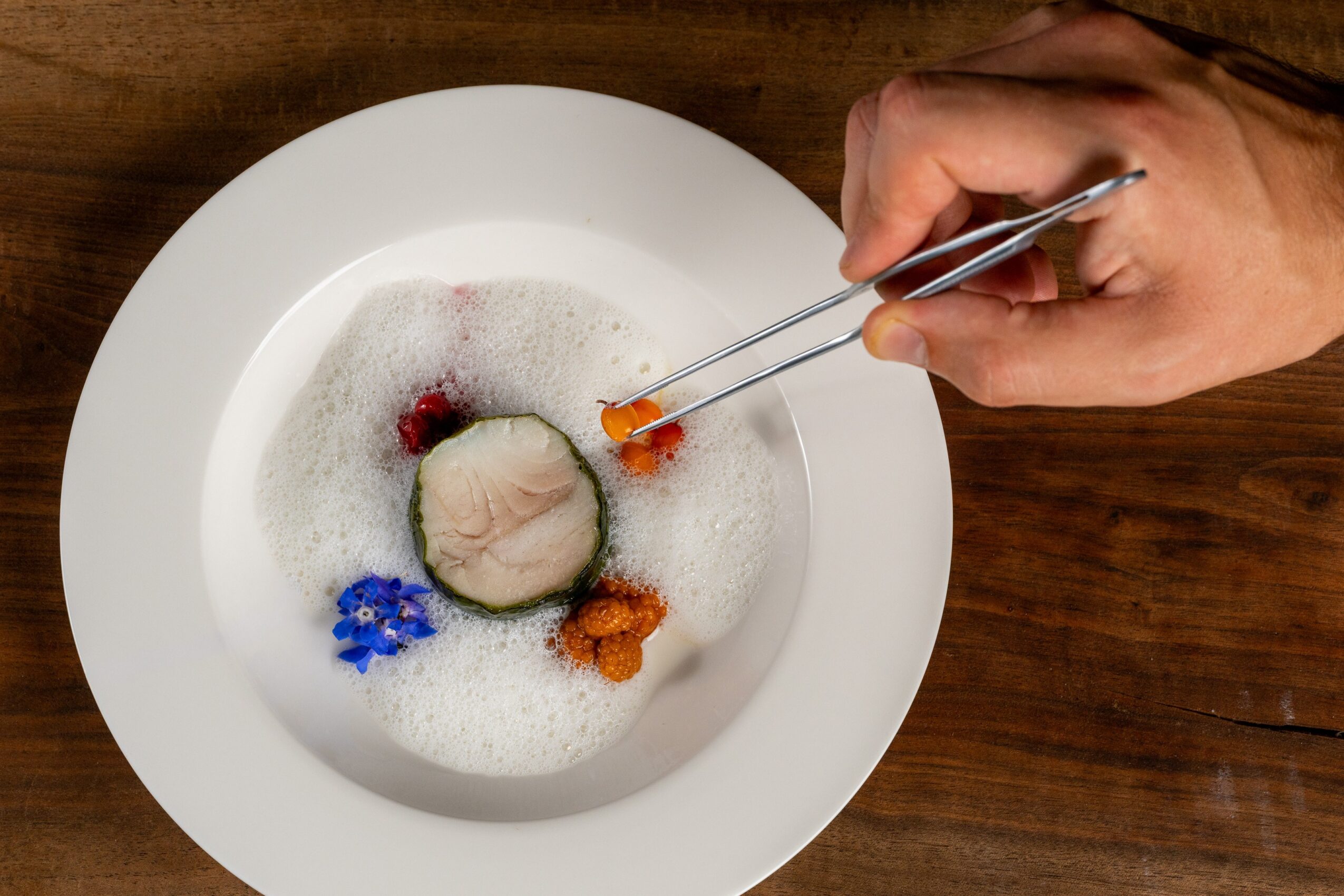 Person using tweezers to place a small orange item into a dish with foam, berries, and edible flowers on a wooden table.