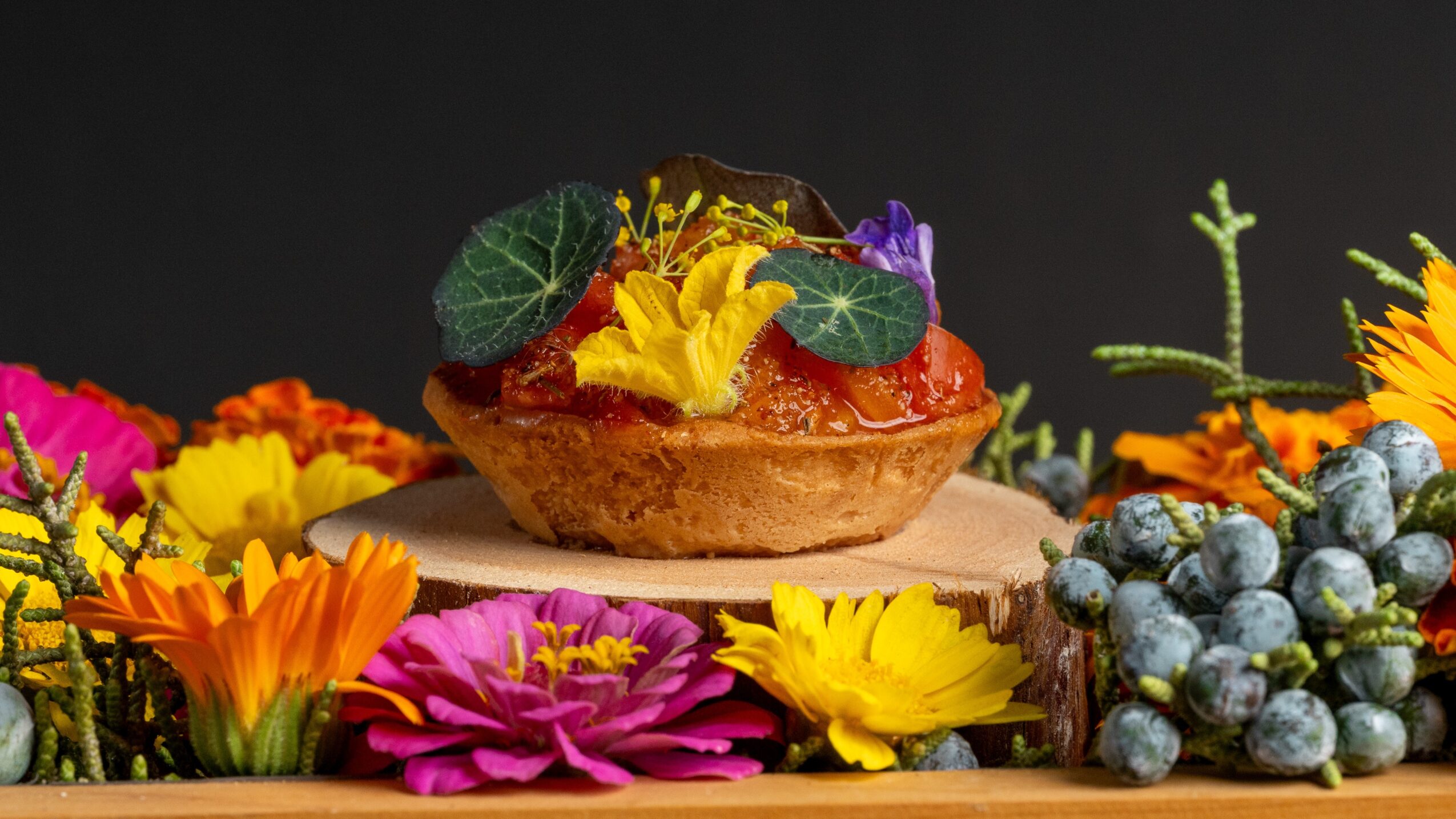 A decorated pastry with edible flowers, leaves, and berries, surrounded by colorful flowers and berries on a wooden surface.