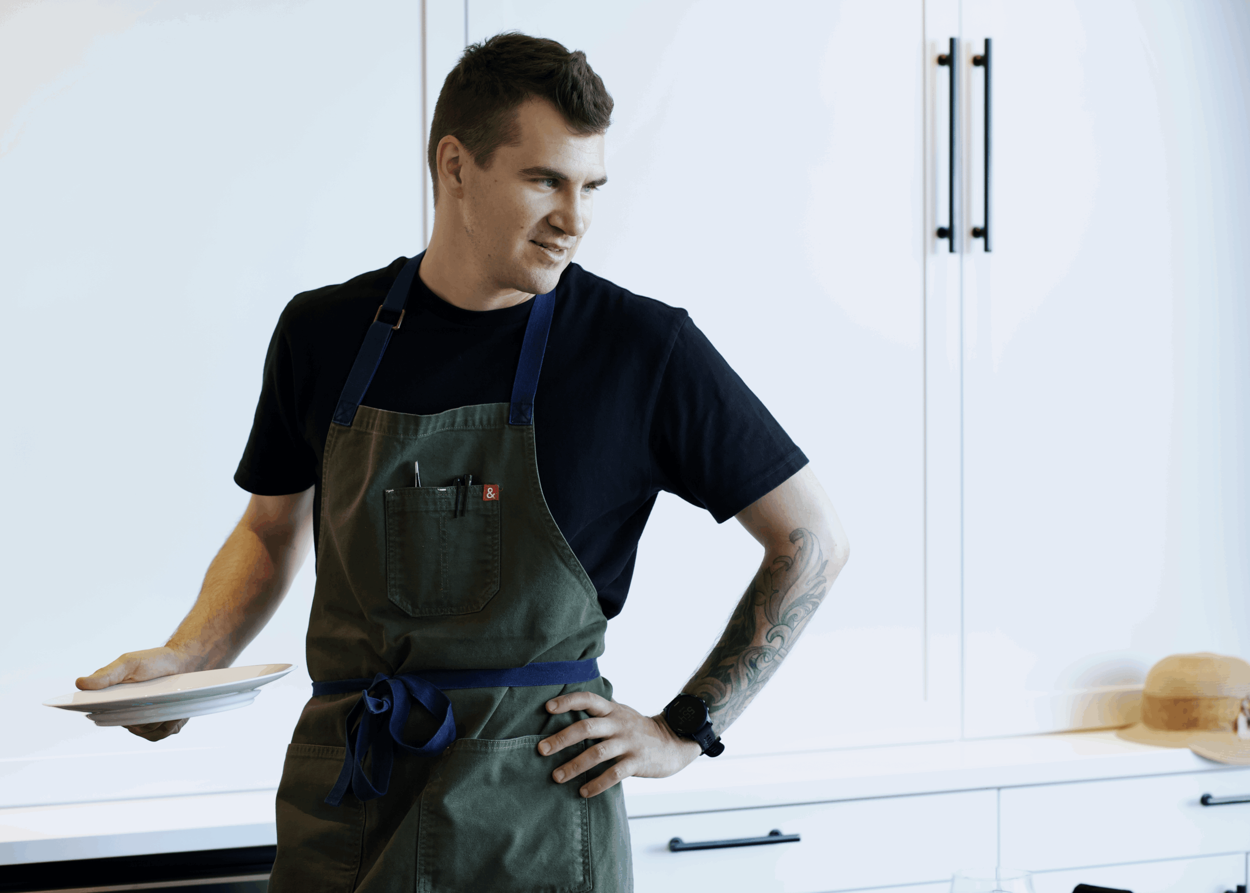 Young man in black shirt and apron standing in kitchen, holding a plate, with cabinets and a cutting board in background.