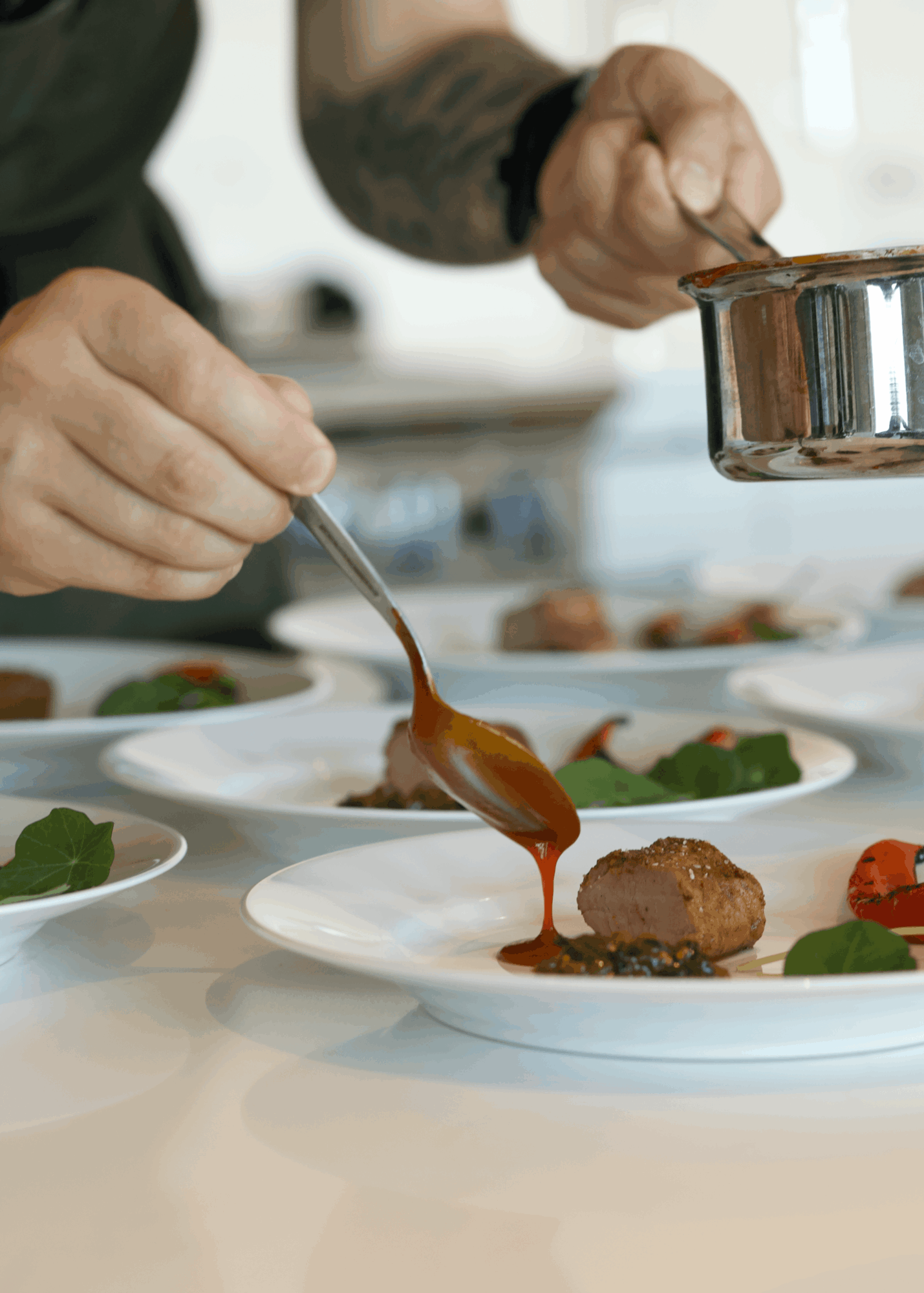 Chef drizzling sauce onto plated food in a professional kitchen, with multiple dishes on the counter.