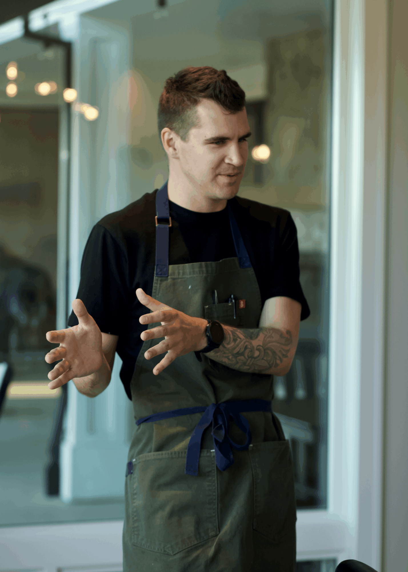 Young man with short hair wearing an apron, gesturing with hands, standing indoors near a window.