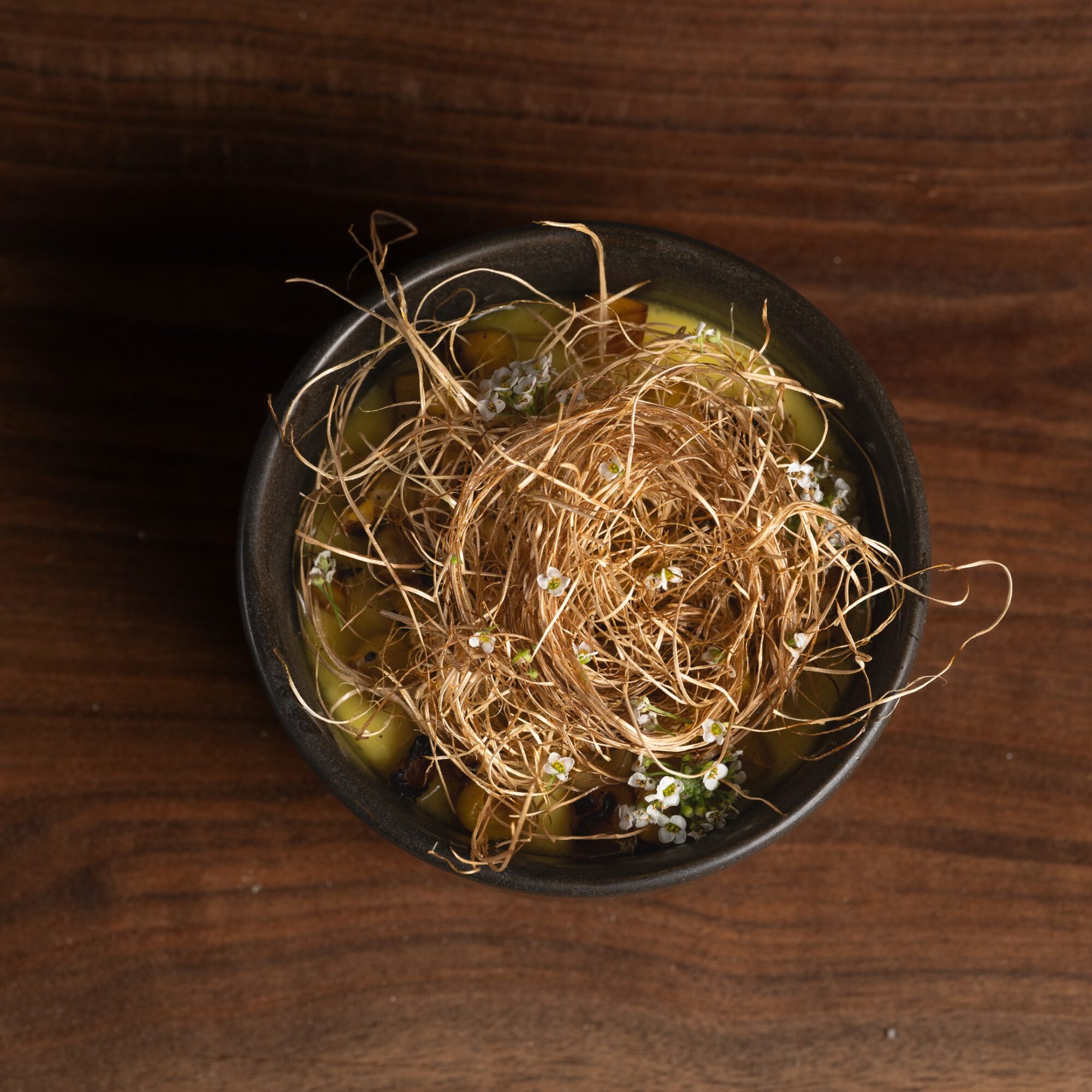 Top-down view of a small black bowl with tangled plant roots and green seeds on a wooden surface.