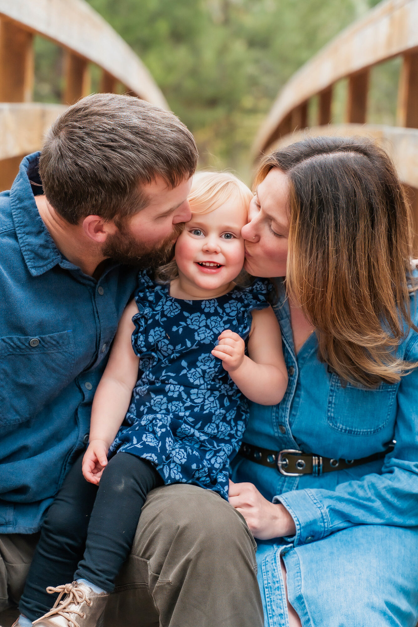 A family of three, a man, woman, and young girl, sitting on a wooden bridge outdoors, smiling and close together.