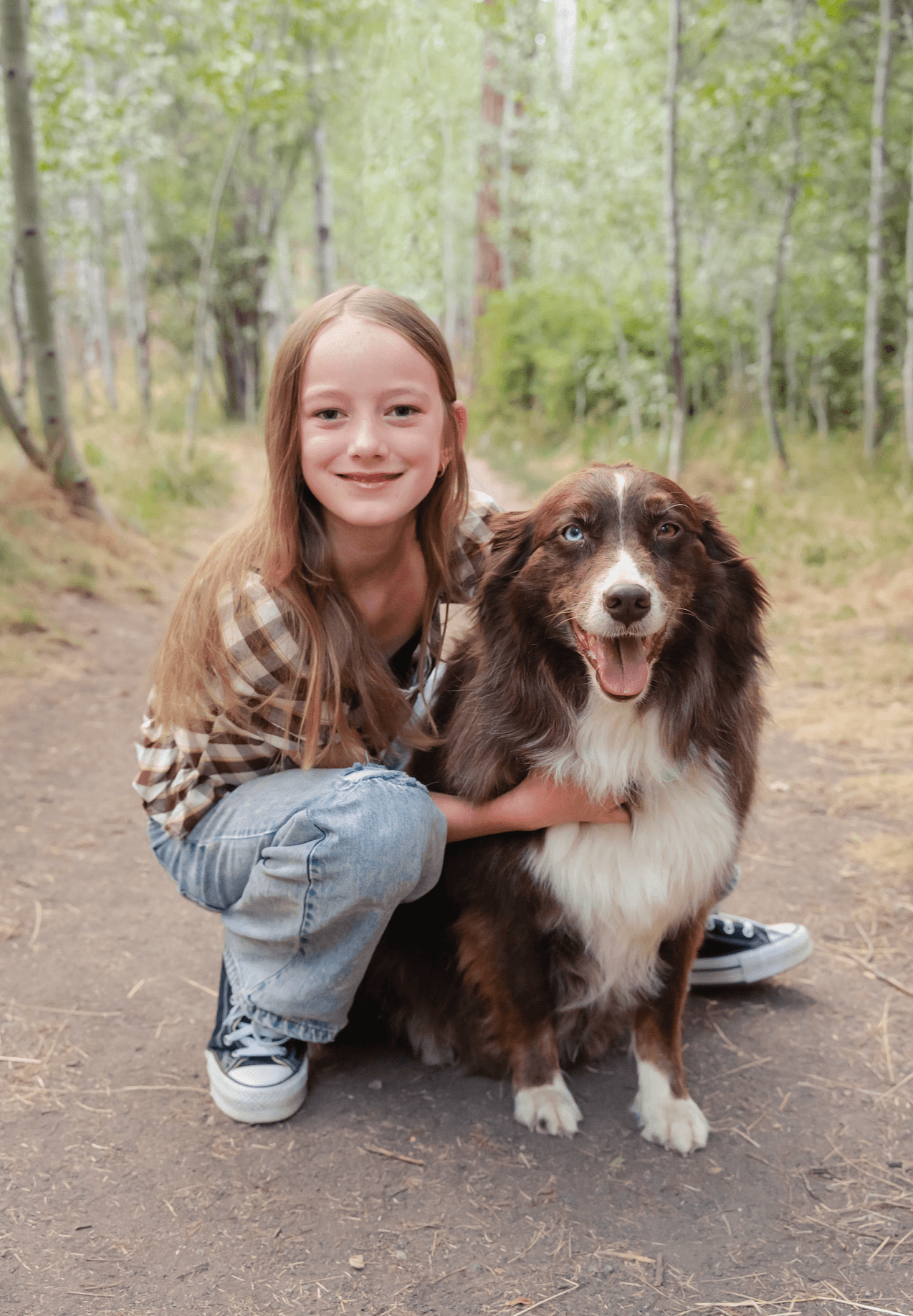 Girl crouching with a dog on a forest trail, smiling, surrounded by trees and greenery.