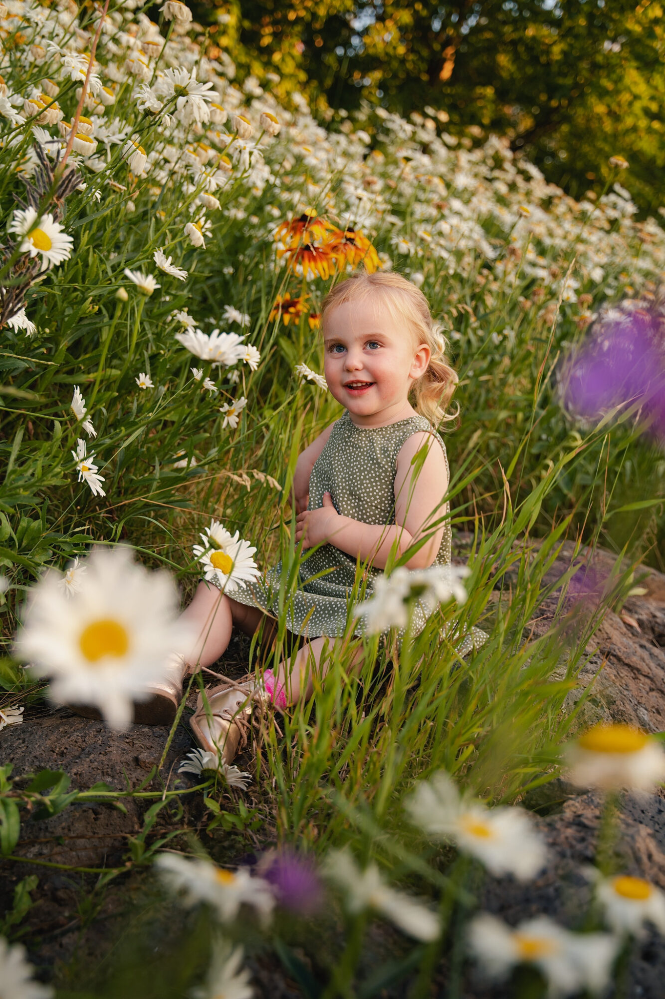 Young girl with blonde hair smiling among white daisies and colorful flowers in a garden.