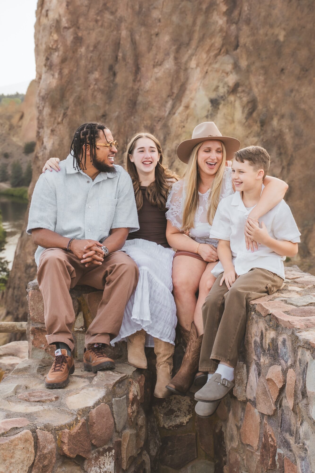Four people sitting on a stone wall outdoors, smiling and embracing each other, with a rocky background.