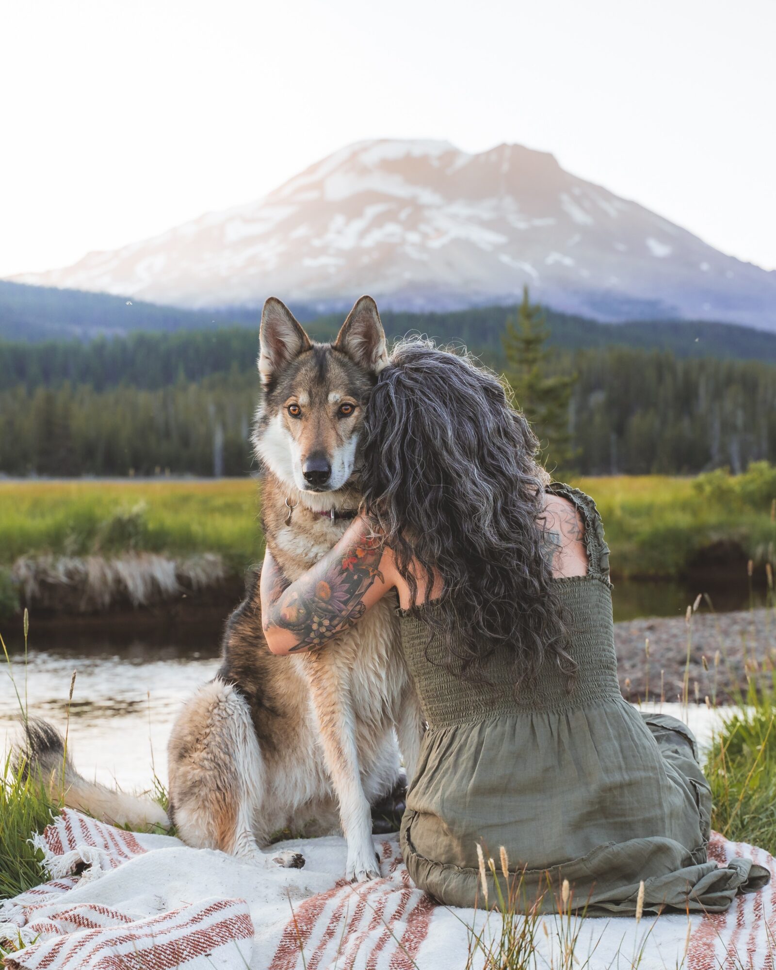 Person with long dark hair hugging a dog outdoors with a mountain and forest in background.