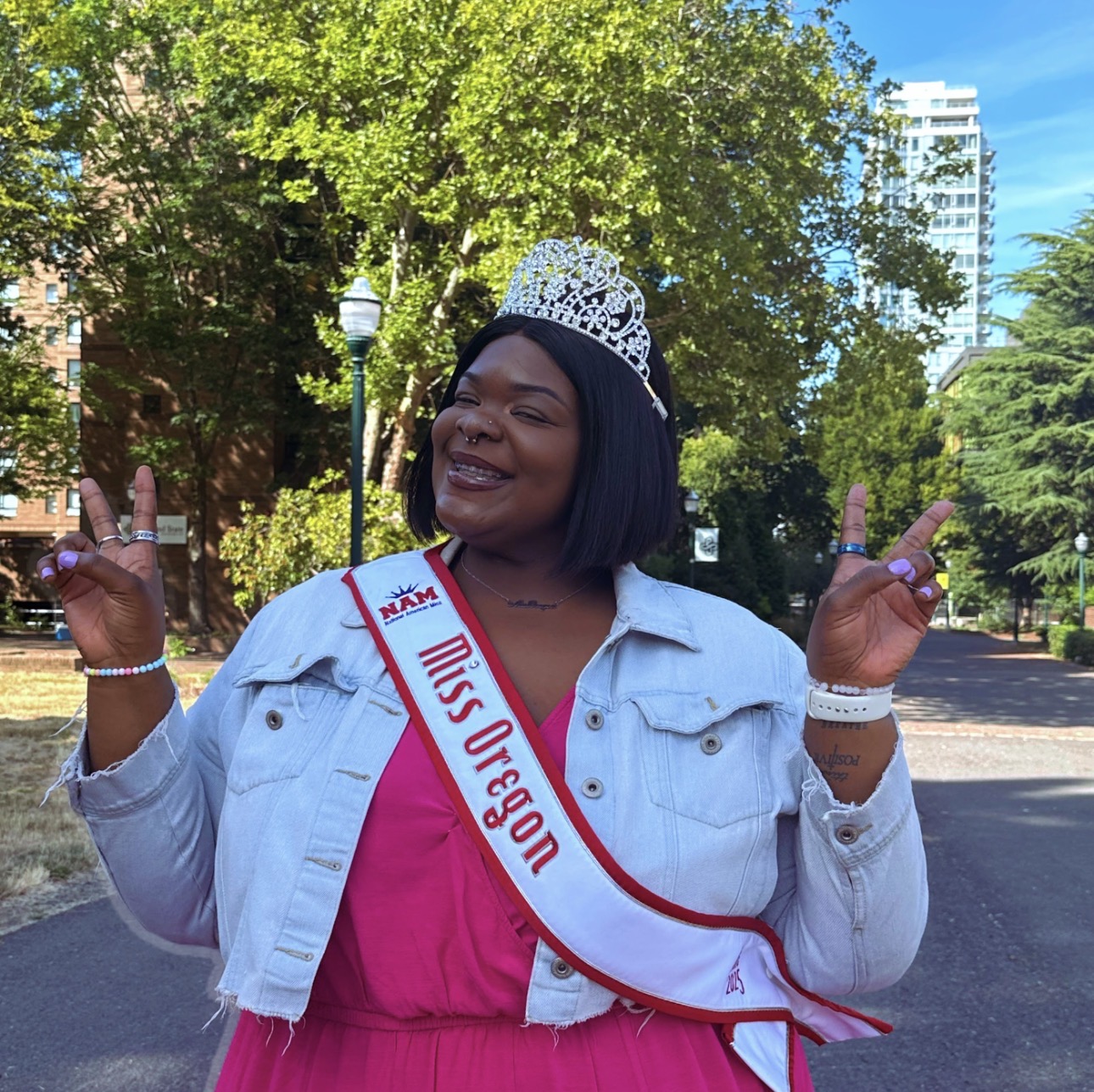 Woman wearing a crown and sash that reads 'Miss Oregon' smiling outdoors with trees and a building in the background.