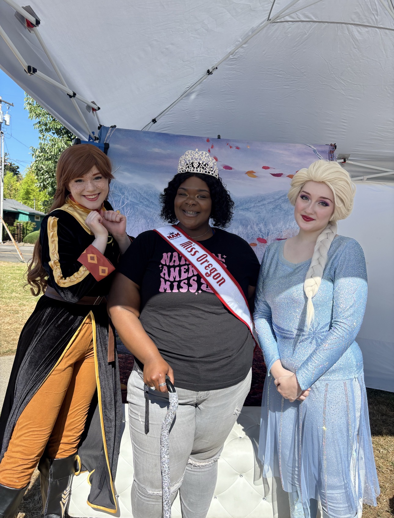 Three women standing under a tent, one wearing a crown and sash, another in a costume, and the third in a blue dress.