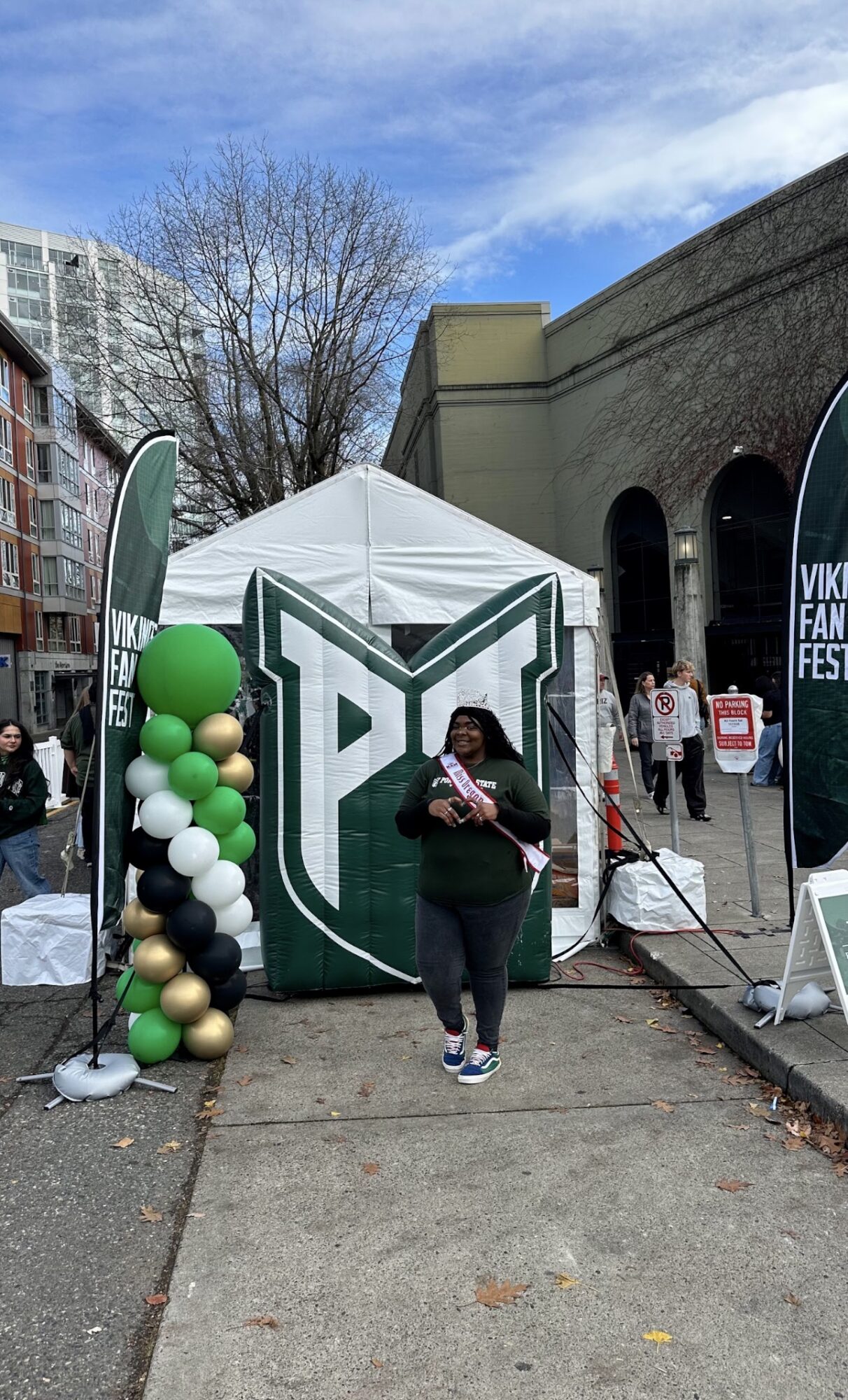 Person standing in front of a tent with green and white balloons, outdoors on a sidewalk, with buildings and trees nearby.