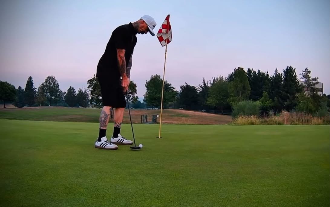 Person preparing to putt on a golf course with trees and a flag in the background.