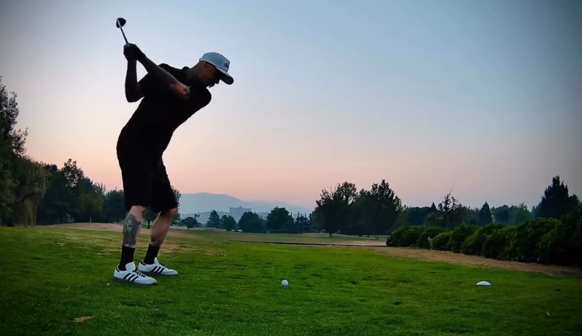 Person in black clothing and cap preparing to swing golf club on a green course at sunset